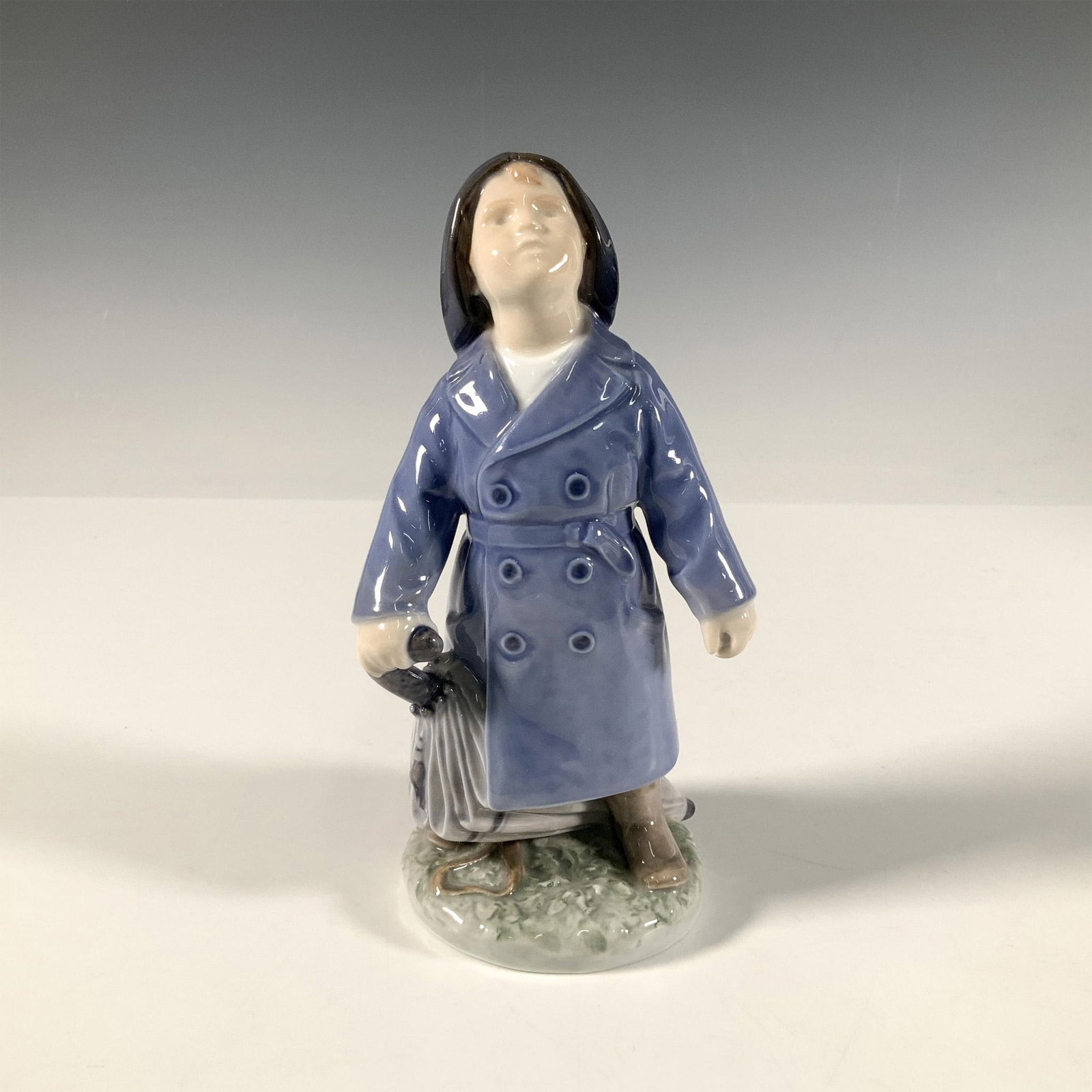 Royal Copenhagen Porcelain Figurine, Boy with Umbrella 3556: A glossy figure of a boy dressed in a blue raincoat and hat, ready for the rainy weather. Royal Copenhagen backstamp. Artist: Adda Bonfils Issued: 20th century Dimensions: 3.5"L x 3"W x 7.25"H Manufac
