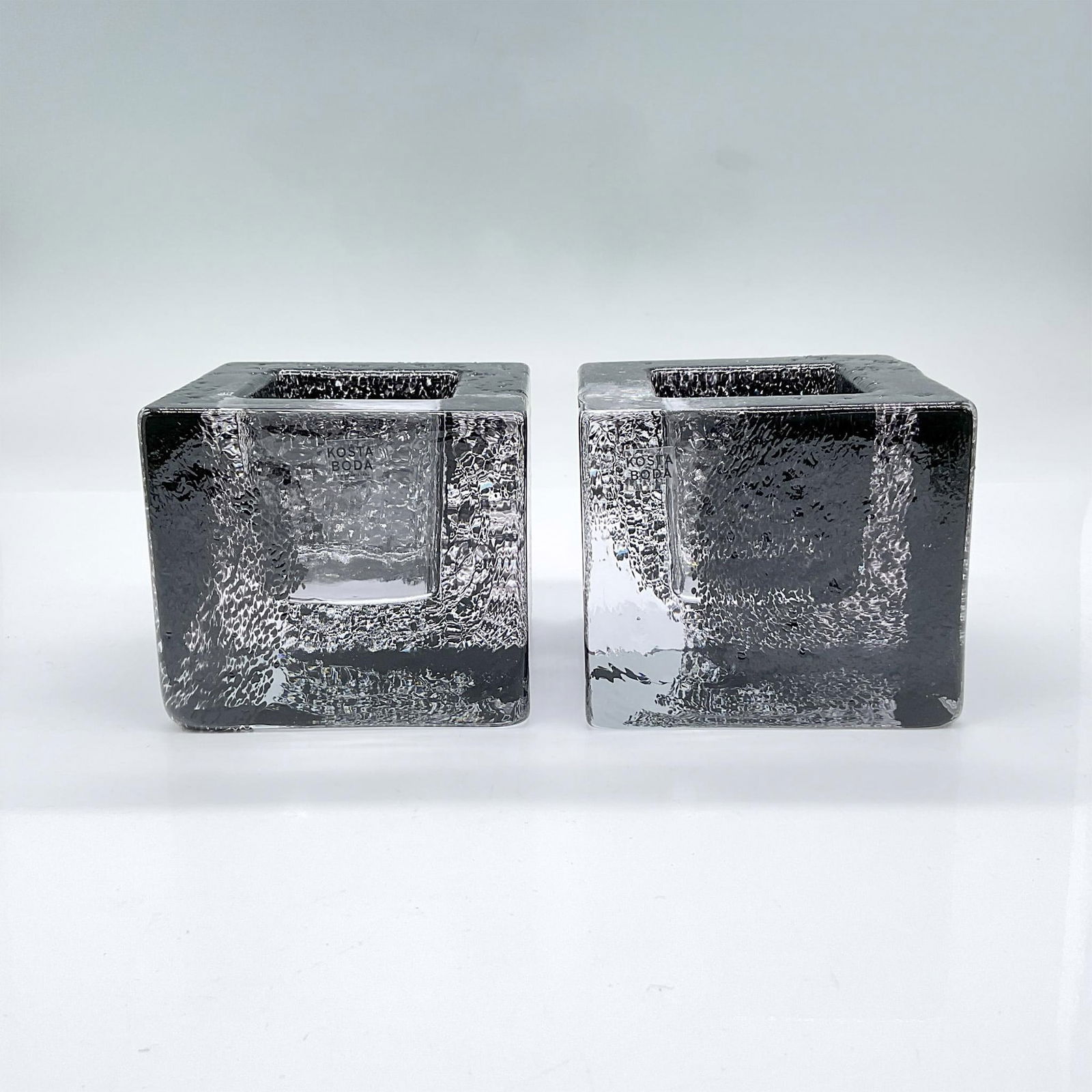 2pc Kosta Boda Art Glass Brick Votives, Spotted Black: Pair of blown and hand formed cube design of clear crystal with a black mottled design. Kosta Boda mark. Includes original box 3.75"L x 3.75"W x 3.25"H. Issued: c. 2005 Dimensions: 3.25"L x 3.25"W x 3