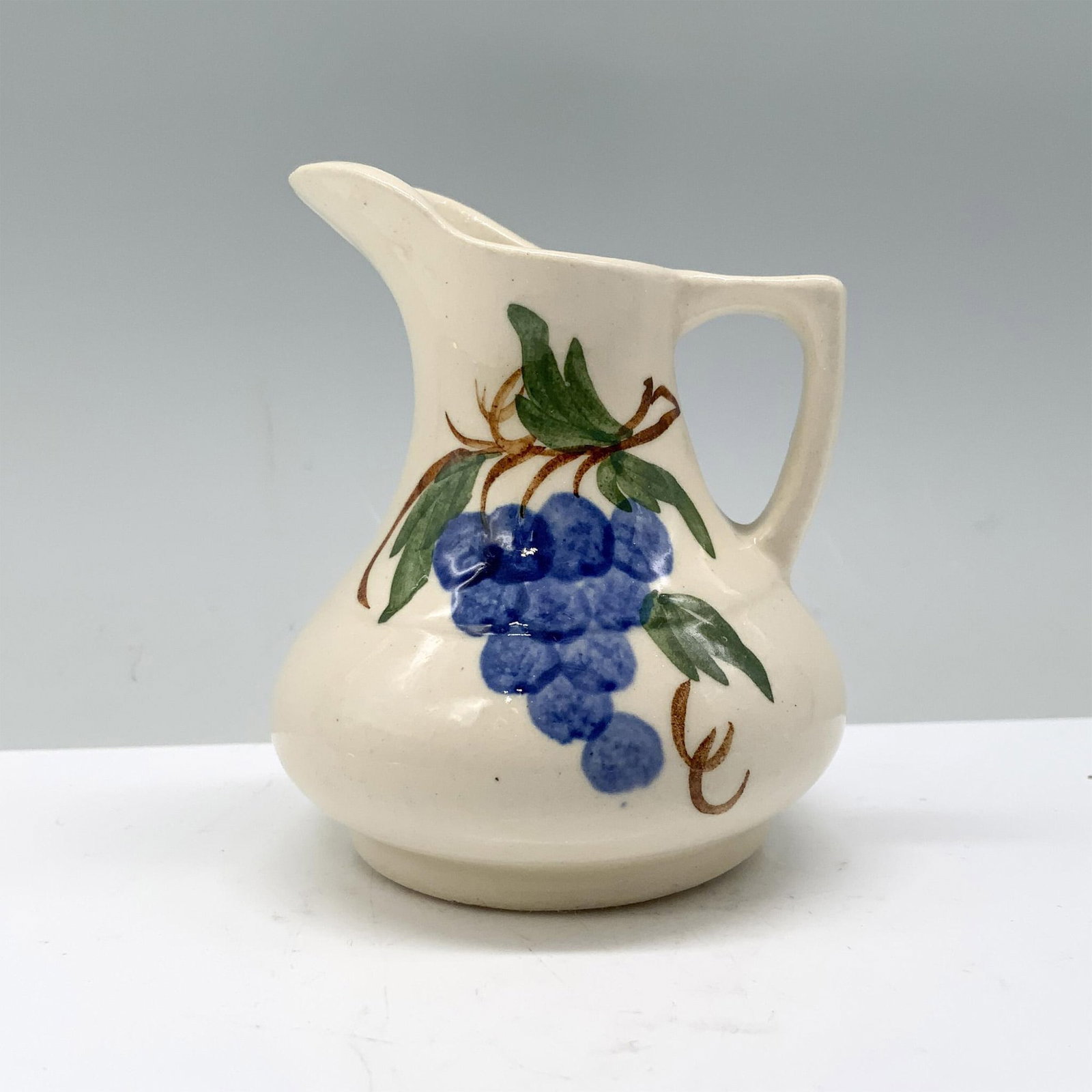 Cash Family Art Pottery Creamer, Blueberries: Cream-colored and adorned with exquisite artwork depicting a cluster of plump blueberries on their branch. Marked Cash Family. Dimensions: 2.5"L x 2.5"W x 3"H Manufacturer: Cash Family Country of Orig