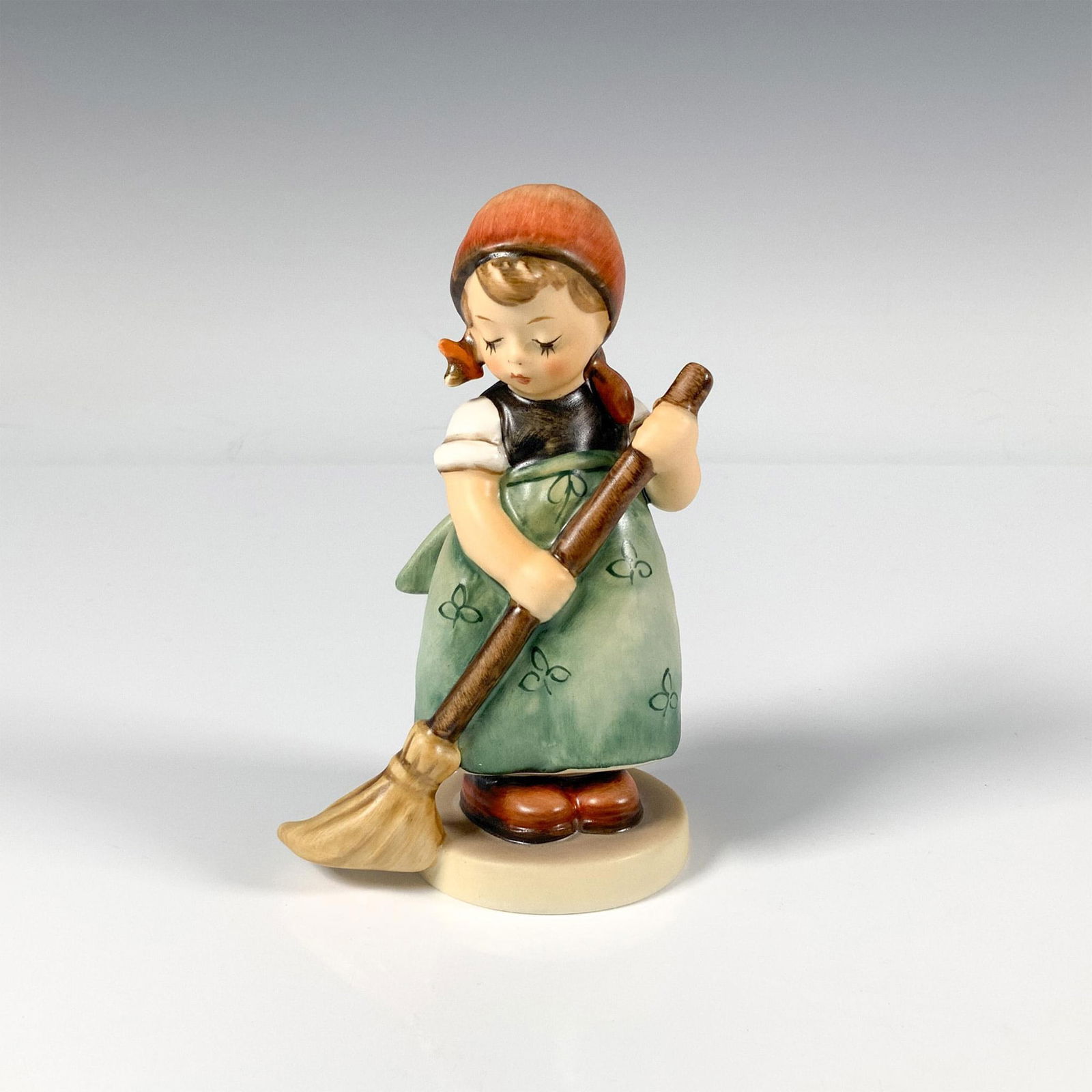 Goebel Hummel Porcelain Figurine, Little Sweeper 171: A small figure of a girl wearing a green apron decorated with a delicate floral pattern, a red scarf around her head and is holding a broom as she sweeps. Goebel backstamp. Artist: M.I. Hummel Issued: