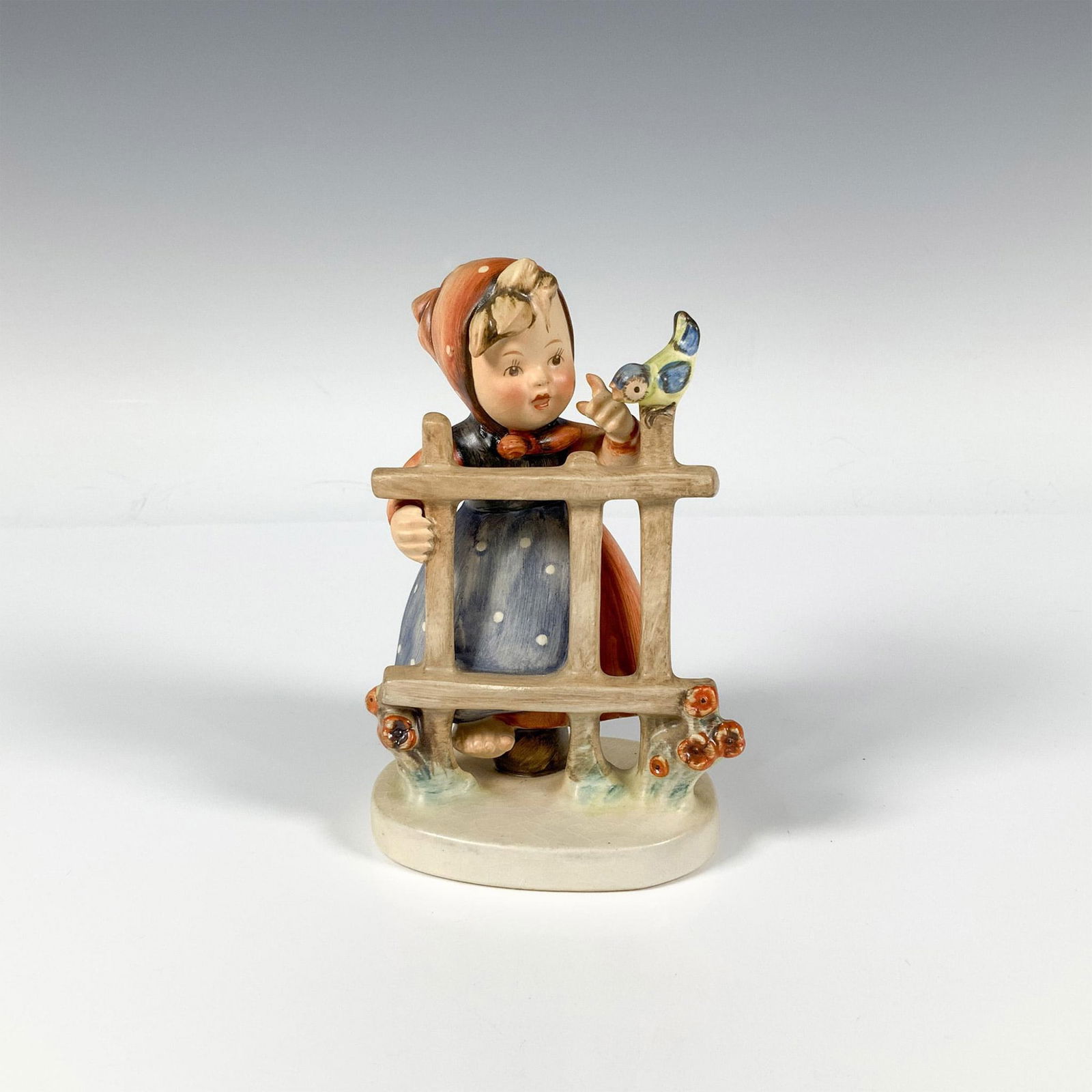 Goebel Hummel Porcelain Figurine, Signs Of Spring 203: A figure of a girl standing by a fence with bird wearing a red dress and scarf around her head. M.I. Hummel impressed. Goebel backstamp. Issued: 1948-1990 Dimensions: 3.25"L x 3"W x 5.5"H Manufacturer