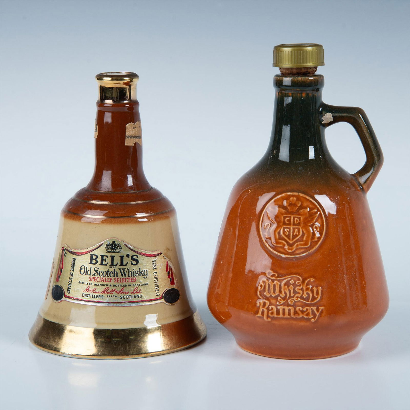 2pc Ceramic Whisky Bottles, Bell's & Whisky Ramsay: The Bell's Old Scotch Whisky bottle features a bell shape in brown and mustard coloration, adorned with gold accents, measuring 7.5"H x 4.75" dia. Ramsay Whisky, of a similar shape with a thumb handle