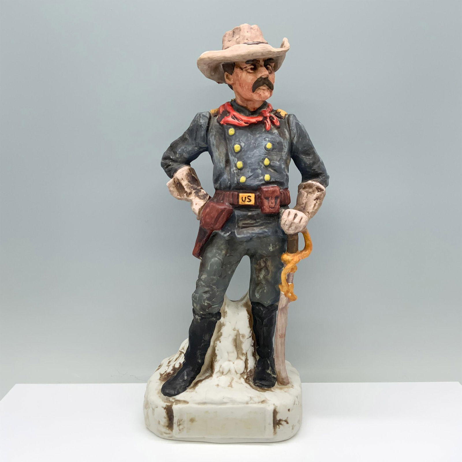 Alpha Industries Decanter by Gary Schildt, Major Reno: A porcelain decanter depicting a cowboy in a blue suit, brown hat, and red ascot, standing resolutely with his saber at his side. Alpha Industries backstamp. Issued: 1975 Dimensions: 5.5"L x 3.75"W x