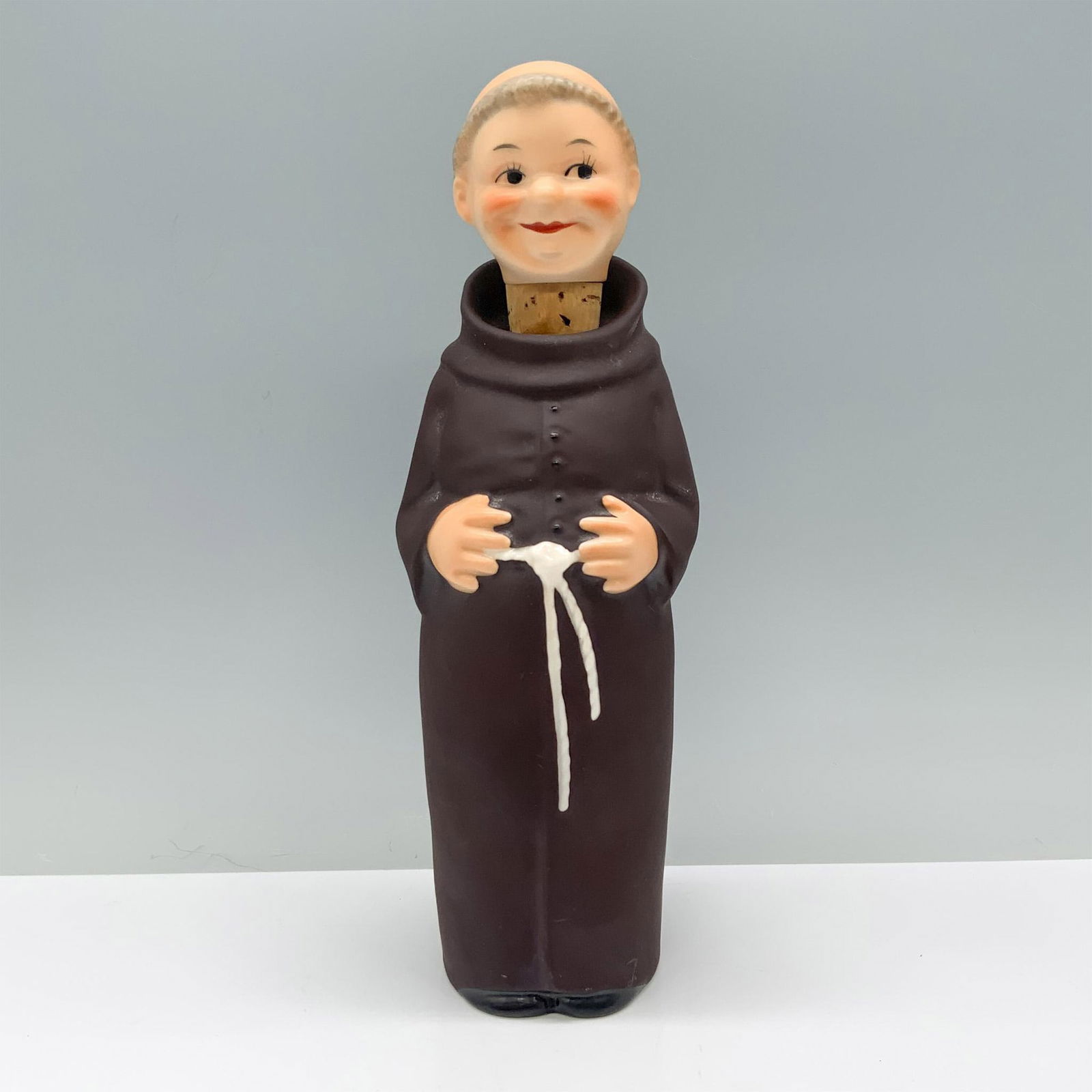 Goebel Porcelain Decanter, Friar Tuck: Friar Tuck, clad in a brown robe with a white belt rope, holds his hands gently against his stomach. Goebel backstamp. Marked KL95. Issued: 1956 Dimensions: 3.5"L x 2.75"W x 10"H Manufacturer: Goebel