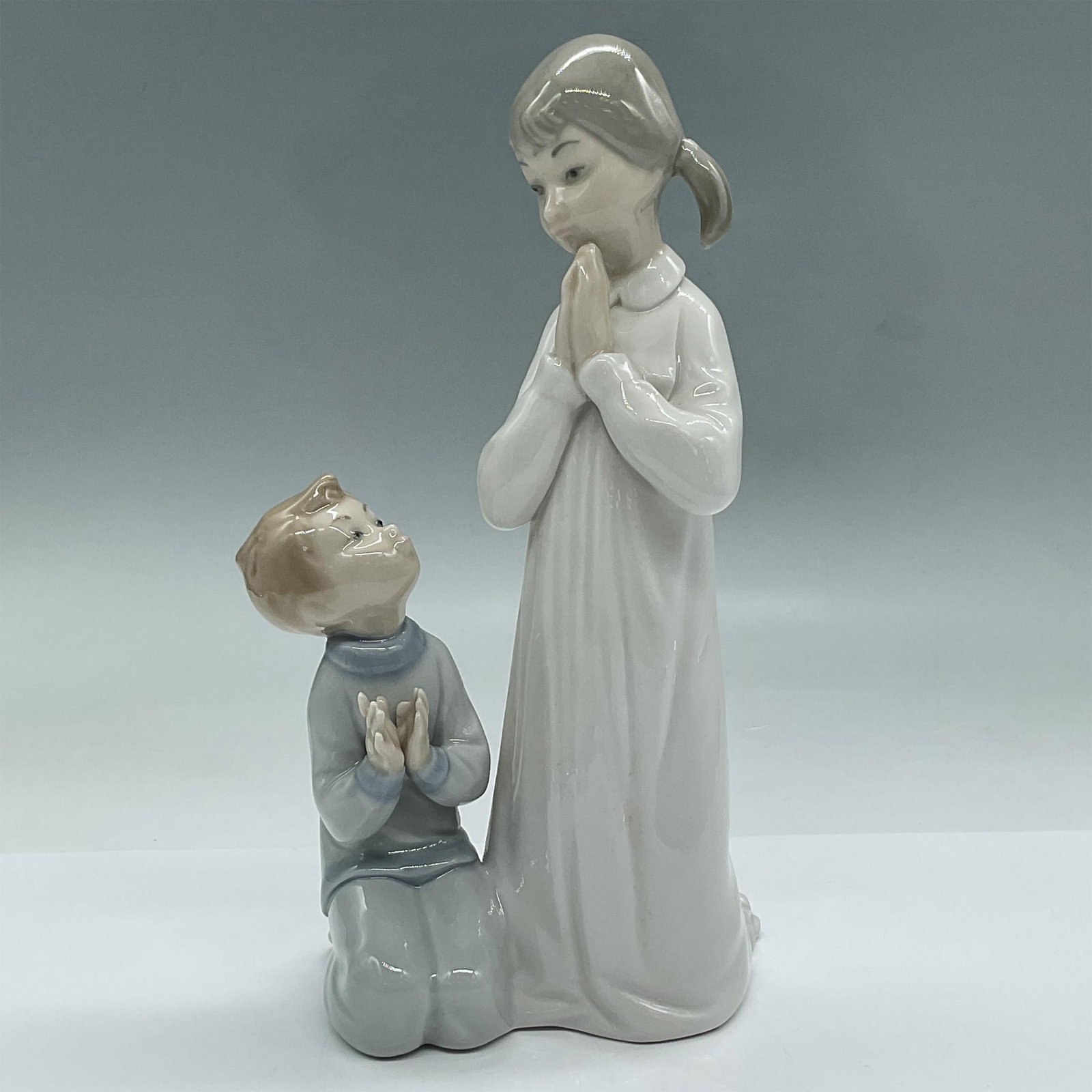 Teaching to Pray 1004779 - Lladro Porcelain Figurine: This artwork beautifully depicts an older sister showing her little brother how to pray. Both dressed and ready for bed. Lladro backstamp. Issued: 20th c. Dimensions: 4"L x 3.5"W x 8.75"H Manufacturer