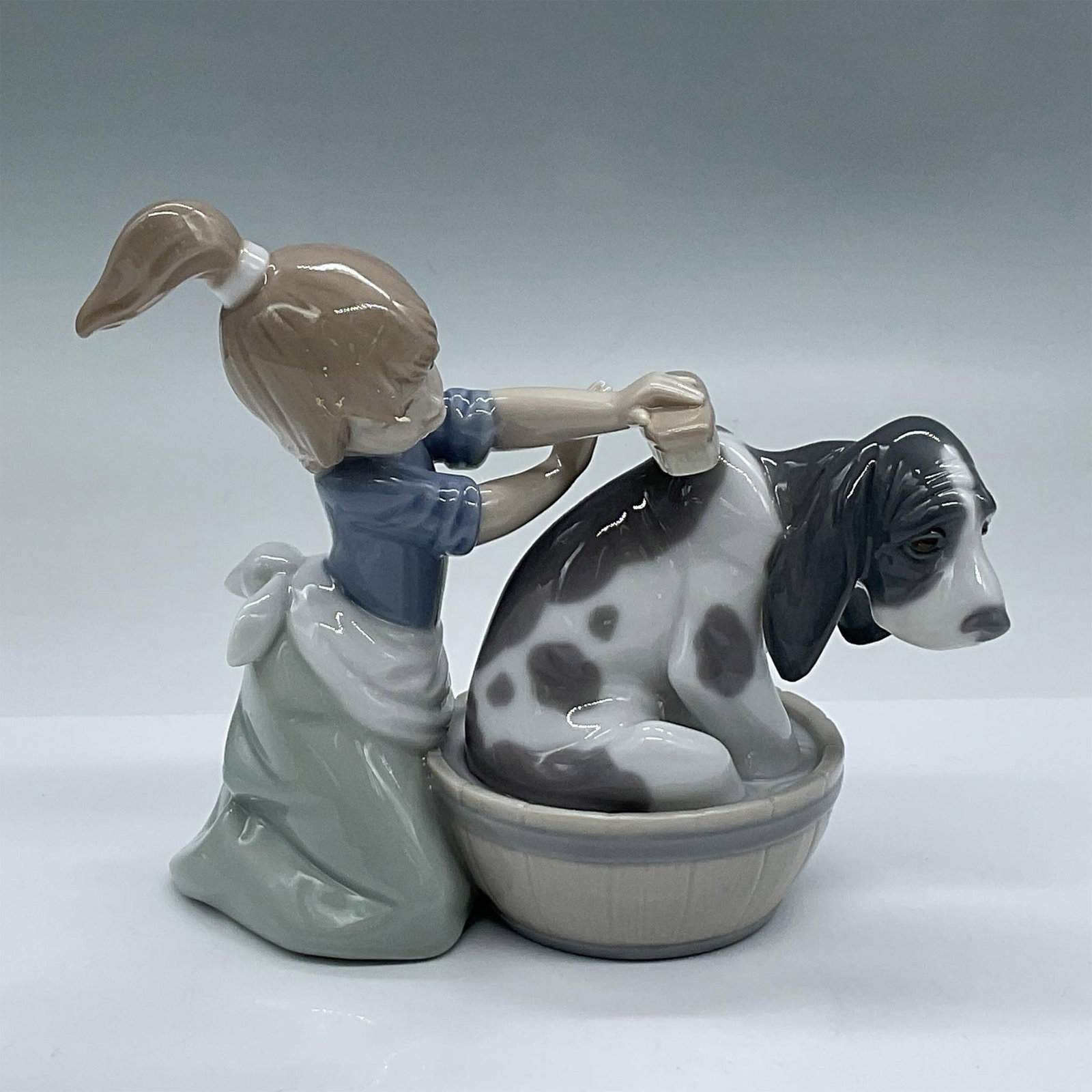 Bashful Bather 1005455 - Lladro Porcelain Figurine: Depicts in soft pastels with a glossy finish. A girl with a ponytail bathing her large pup in a small bucket. Lladro backstamp. This item has its original box. Measures 7.25"L x 4.5"W x 7.75"H. Issued