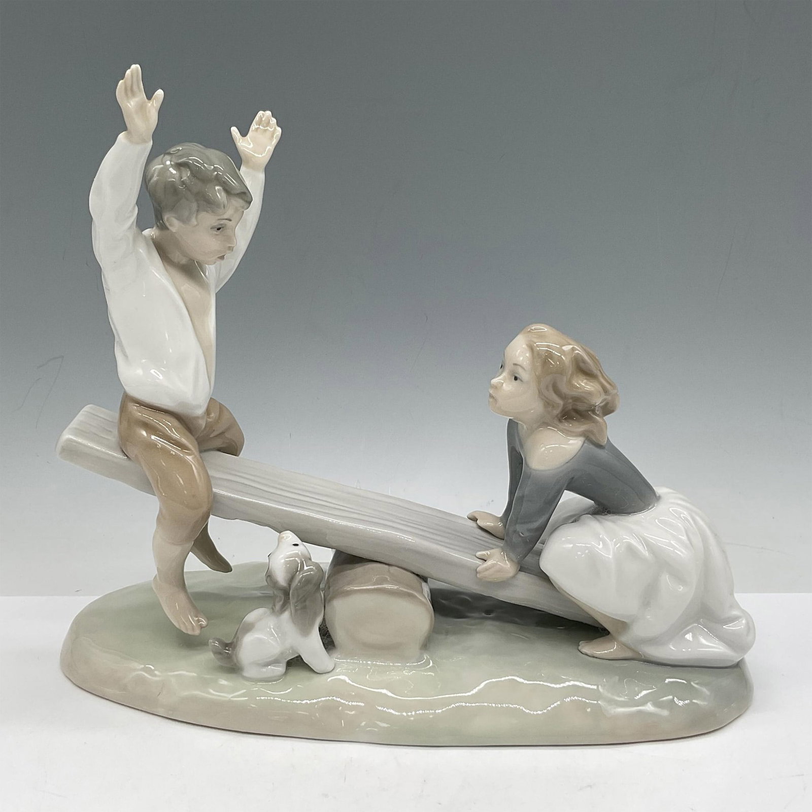 See-Saw 1004867 - Lladro Porcelain Figurine: Children playing on see-saw with dog looking on. Colored in cream, white and gray with glossy finish. Lladro backstamp. Artist: Fulgencio Garcia Issued: 1974 - 1996 Dimensions: 9"L x 4"W x 8"H Manufac