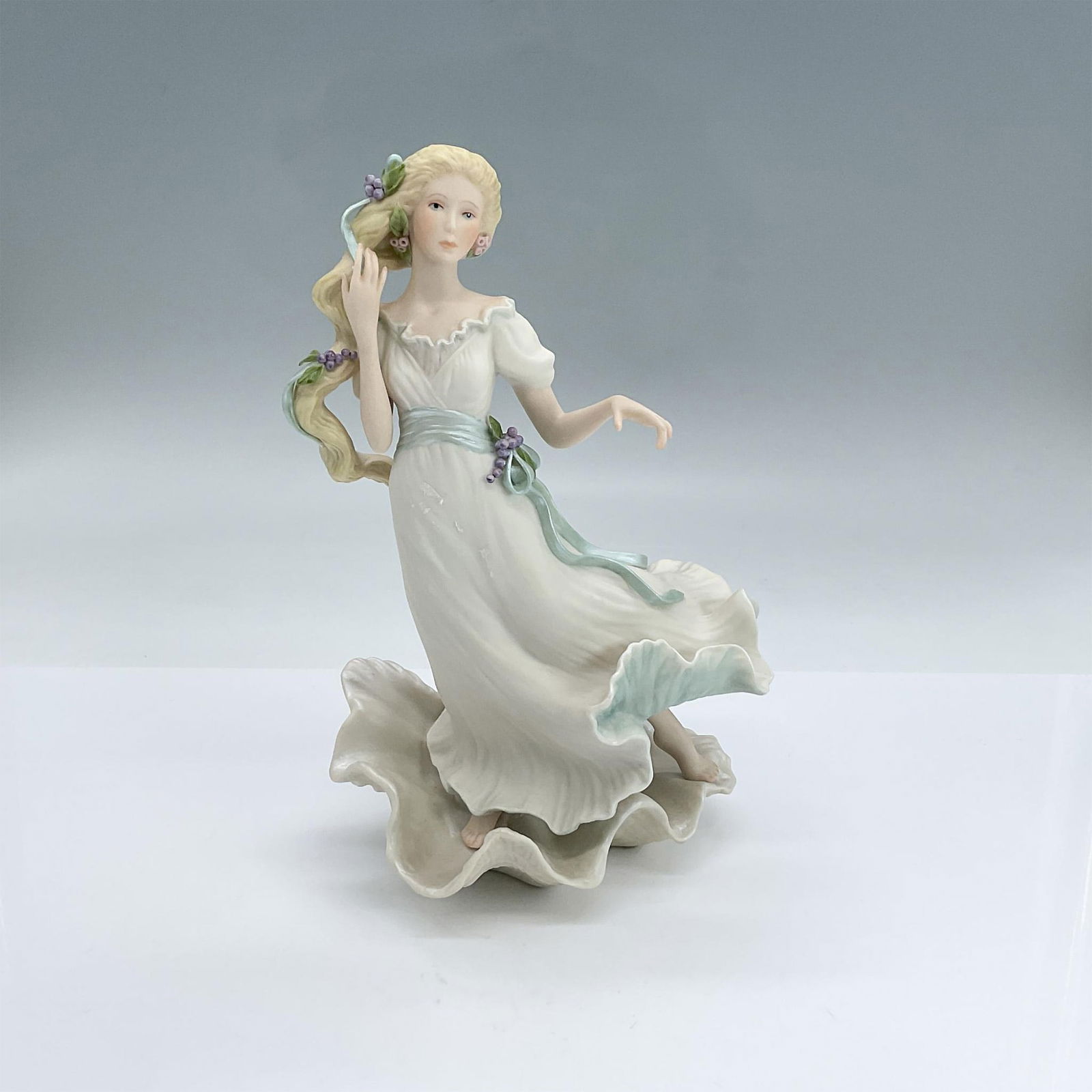 Cybis Porcelain Figurine, Aphrodite: Matte porcelain figure of a lady in a white dress standing on a seashell with purple grapes and blue accents. Cybis backstamp. Dimensions: 5.5"L x 4"W x 8.75"H Manufacturer: Cybis Country of Origin: U