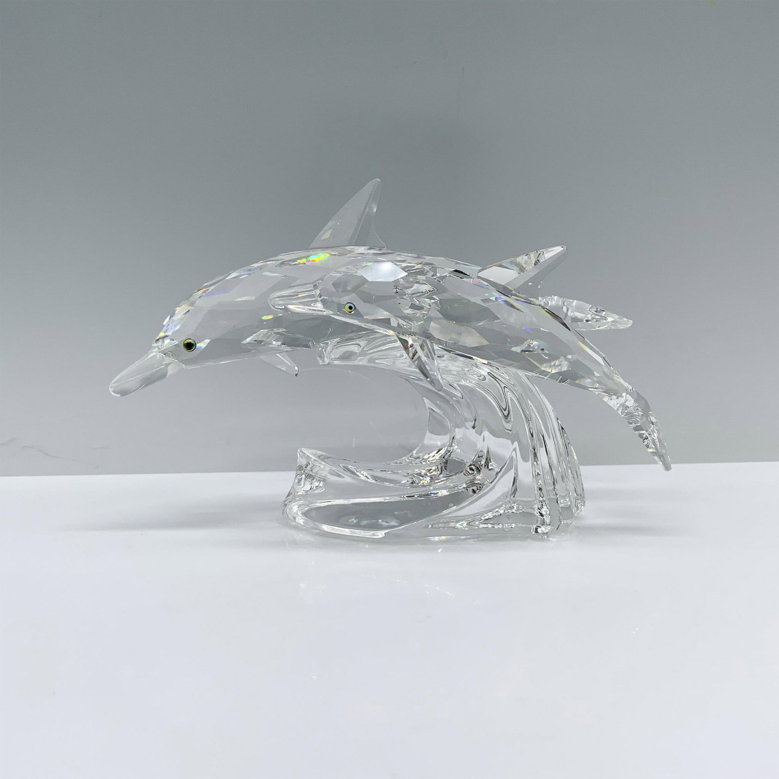 Swarovski 1990 Edition Crystal Figurine, Lead Me 153850: A clear crystal sculpture depicting a mother and child dolphin swimming gracefully side by side over a wave, their eyes adorned in a soothing melon color. Swarovski backstamp. This item has its origin