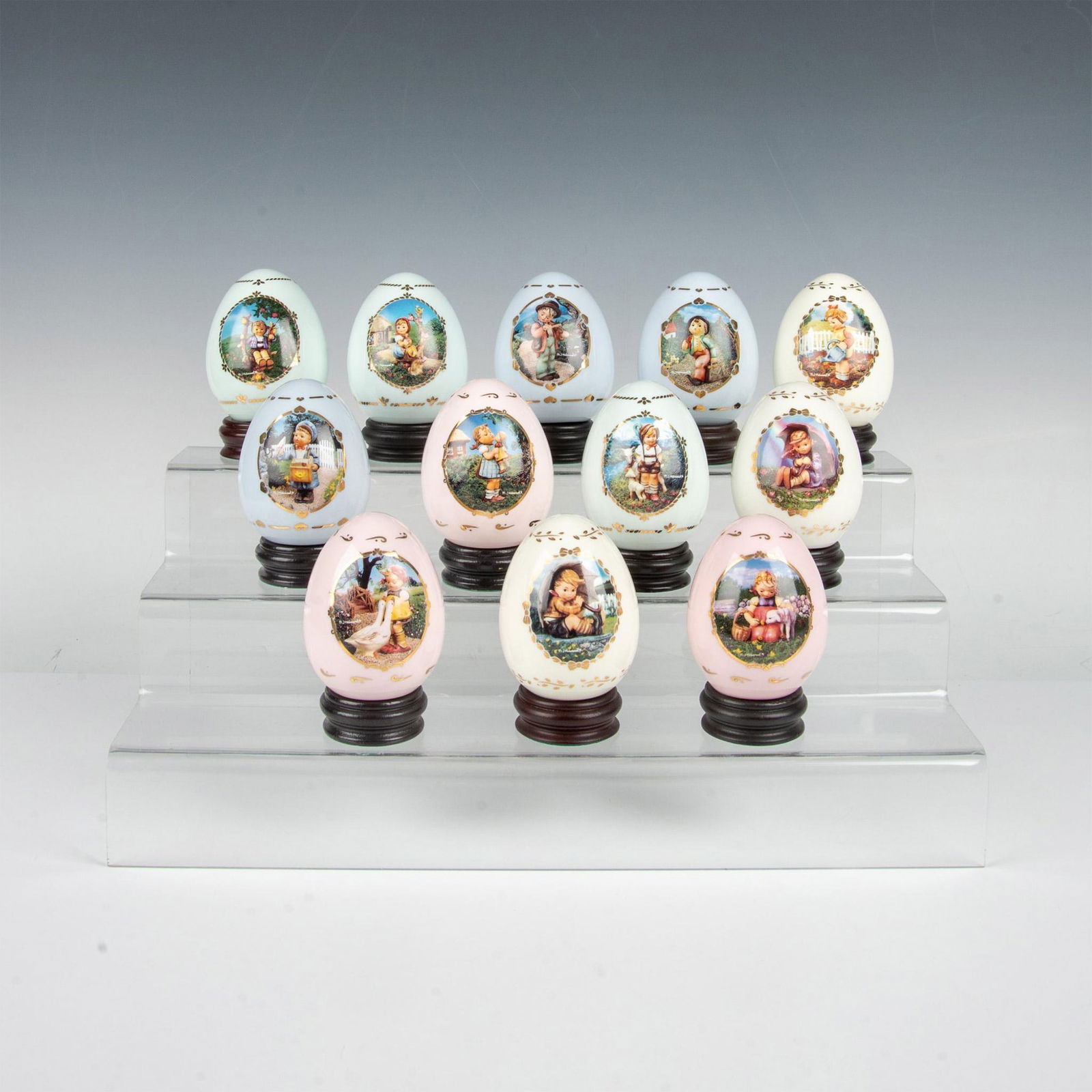 12pc Goebel Hummel Gilded Porcelain Eggs with Bases: Beautiful collection of delightful eggs that capture the essence of the beloved Hummel children in colorful scenes, adorned with gold accents featuring lovely pastel colors. Each is accompanied by its
