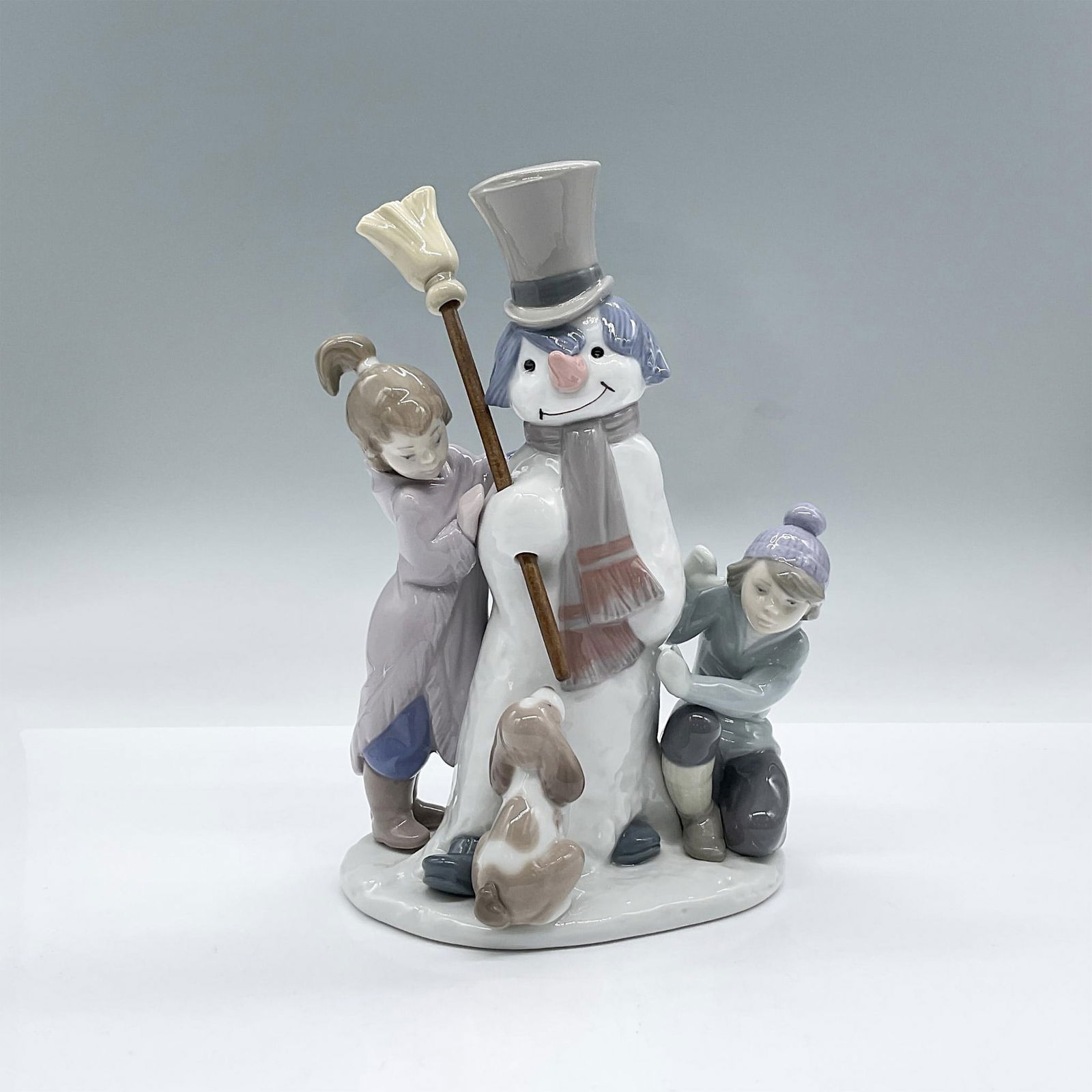 The Snow Man 5713 - Lladro Porcelain Figurine: A glossy figure depicting a winter scene with a couple of children building a snow character with their dog curiously gazing. Lladro backstamp. This item has its original box: 9"L x 6"W x 10"H. Artist