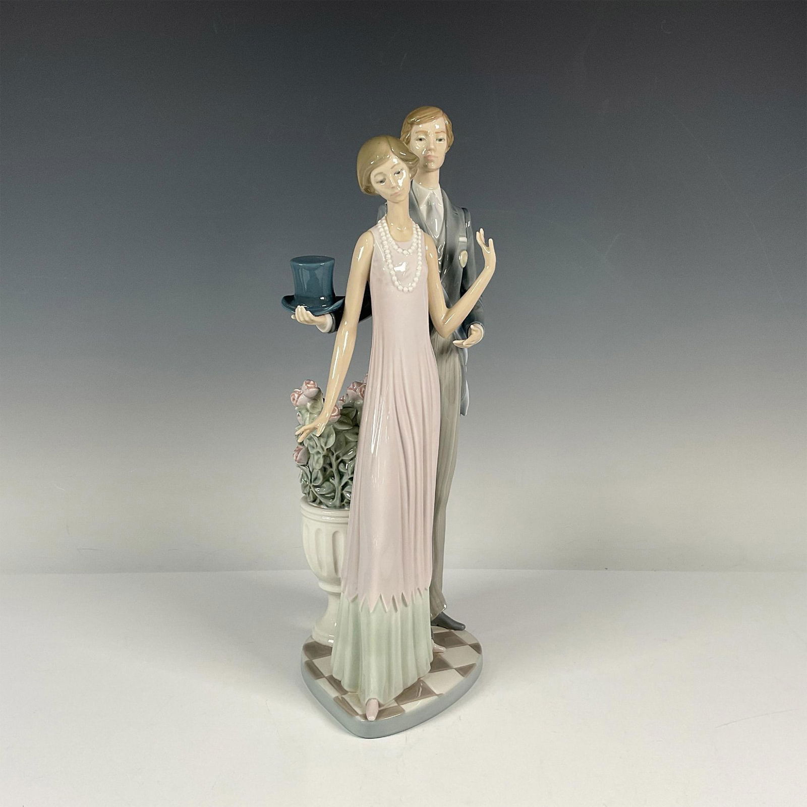 High Society 1001430 - Lladro Porcelain Figurine: Glossy porcelain figure of a finely dressed lady and man standing next to a jardiniere with pink roses. Lladro backstamp. Includes original box: 20"L x 8"W x 7"H. Issued: 1982-1991 Dimensions: 6.5"L x