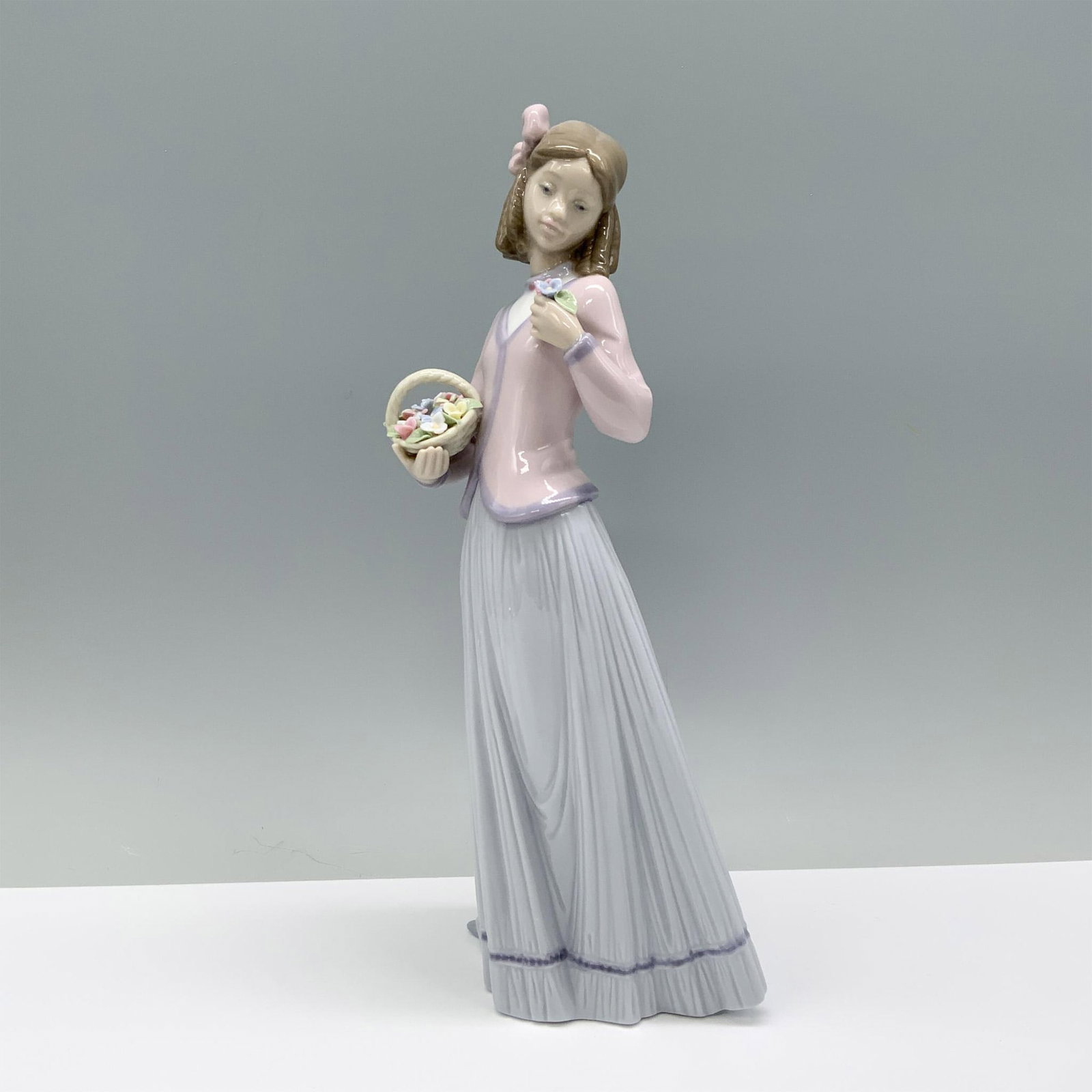 Innocence in Bloom 01007644 - Lladro Porcelain Figurine: A woman elegantly dressed in a pink blouse and lavender skirt gracefully holds a basket of flowers in one hand and a bouquet in the other. Lladro backstamp. This item has its original box which measur