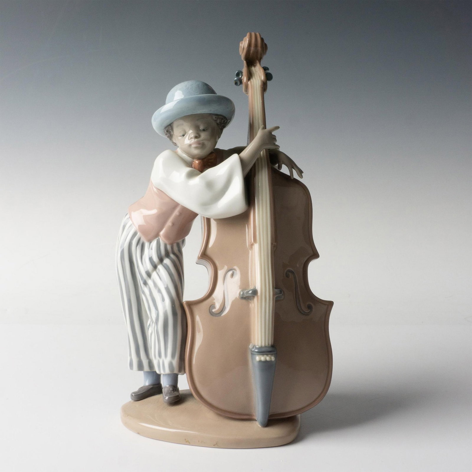 Jazz Bass 5834 - Lladro Porcelain Figurine: A glossy figure of a boy wearing a pink vest and striped pants, with his arm around a bass. Lladro backstamp. Artist: Rafael Lozano Issued: 1991-2017 Dimensions: 5.5"L x 4.25"W x 10"H Manufacturer: Ll