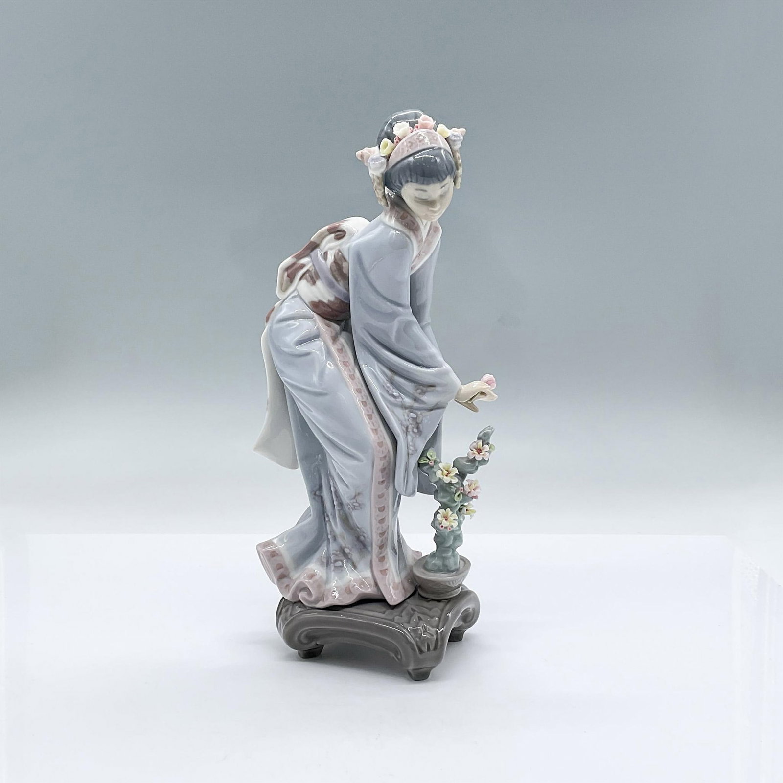 Mayumi 1449 - Lladro Porcelain Figurine: A glossy figure of a woman wearing a kimono with floral designs, bending over a potted plant adorned with flowers. Lladro backstamp. This item has its original box: 12.5"L x 7.5"W x 6"H. Artist: Salva