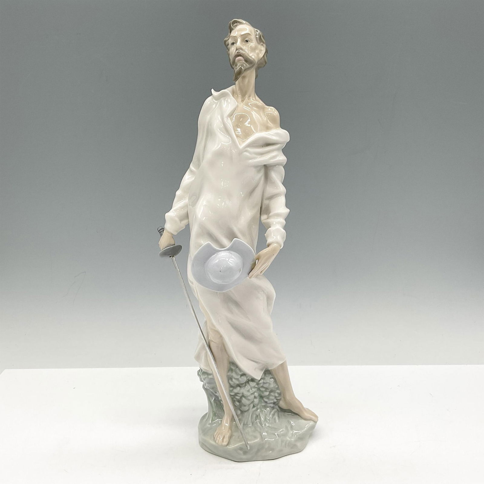 Nao By Lladro Porcelain Figurine, Don Quixote: Glossy figure standing with his sword, hat in hand. Nao backstamp. Issued: 20th c. Dimensions: 3.5"L x 3.5"W x 12"H Manufacturer: Nao by Lladro Country of Origin: Spain