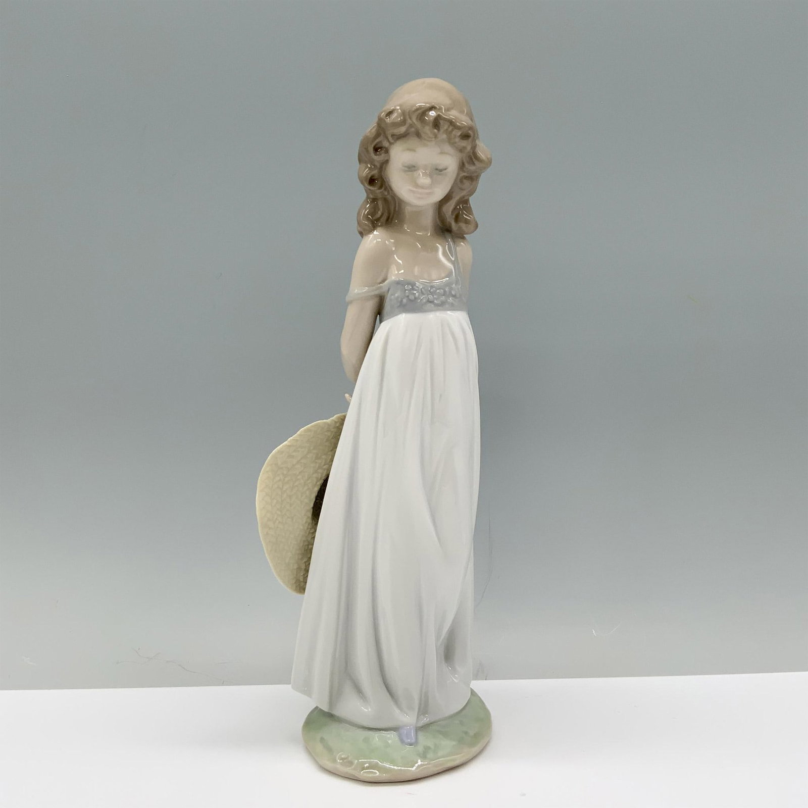 Natural Beauty 01008114 - Lladro Porcelain Figurine: Clad in a white sundress, the girl shyly holds her sunhat behind her back. Lladro backstamp. This item has its original box which measures: 11.25"L x 5.25"W x 5"H. Artist: Antonio Ramos Issued: 2005-2