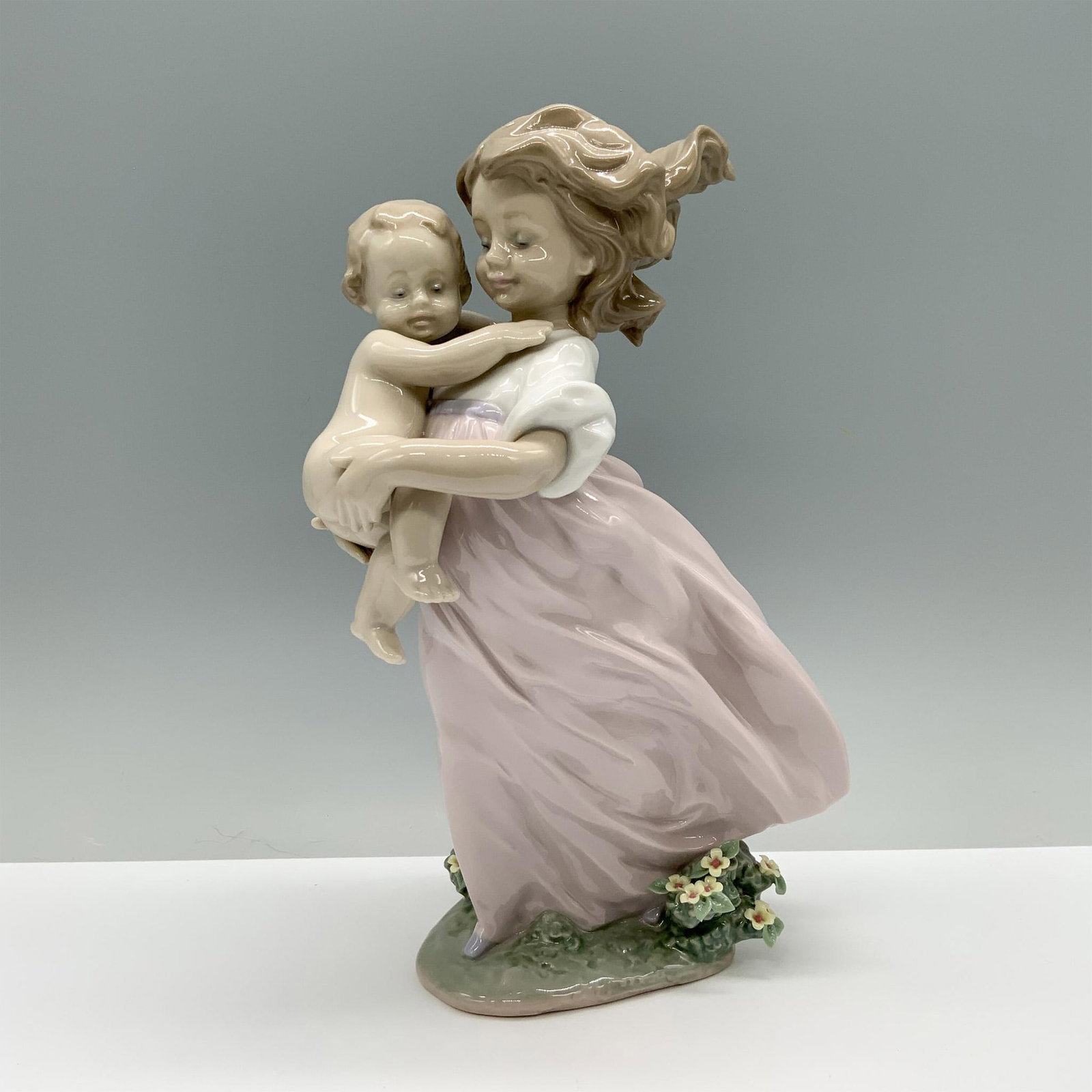 Playing Mom 01006681 - Lladro Porcelain Figurine: A girl in a white shirt and flowing purple skirt embraces her maternal instincts as she gently holds a baby close. Lladro backstamp. This item has its original box which measures: 13"L x 8.25"W x 7"H.