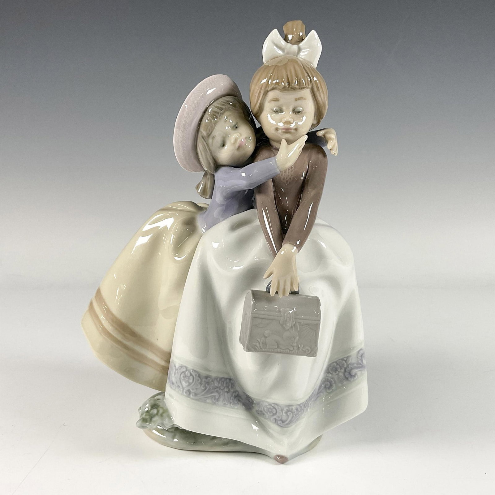 Sharing Secrets 1005720 - Lladro Porcelain Figurine: Glazed finish. Features two adorable girls sharing a loving hug, dressed in prominent dresses. Lladro backstamp. Artist: Antonio Ramos Issued: 1986 - 2023 Dimensions: 5.5''L x 6''W x 8.5''H Manufactur