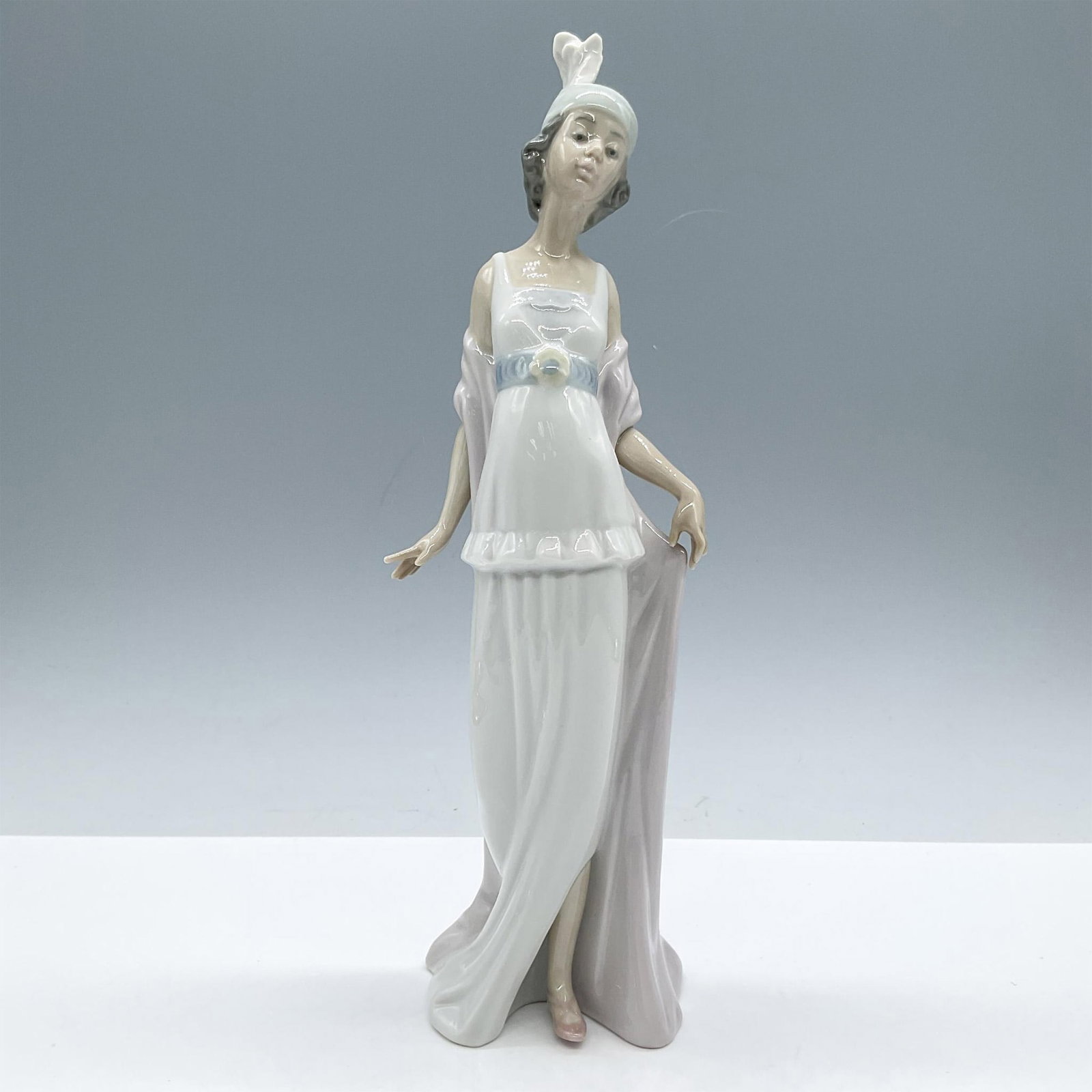 Talk of the Town 1005788 - Lladro Porcelain Figurine: Glossy finish. Depicts an elegant lady, modeled in white blue and pink pale colors. Lladro backstamp. Includes the original box, dimensions: 7.5''H x 12.5''W x 6''H. Artist: Joan Coderch Issued: 1991 