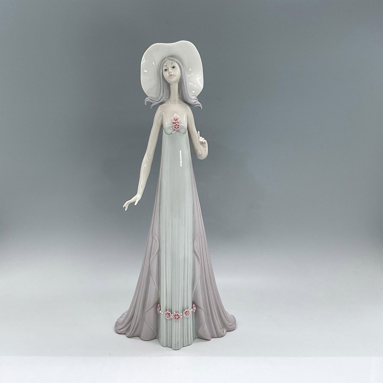 The Debutante 1001431 - Lladro Porcelain Figurine: Glossy porcelain figure of a lady in a long pastel purple and blue dress and white wide-brimmed hat. Lladro backstamp. Includes original box: 7.25"L x 18"W x 7"H. Signed. Dimensions: 6"L x 4.2"W x 13.