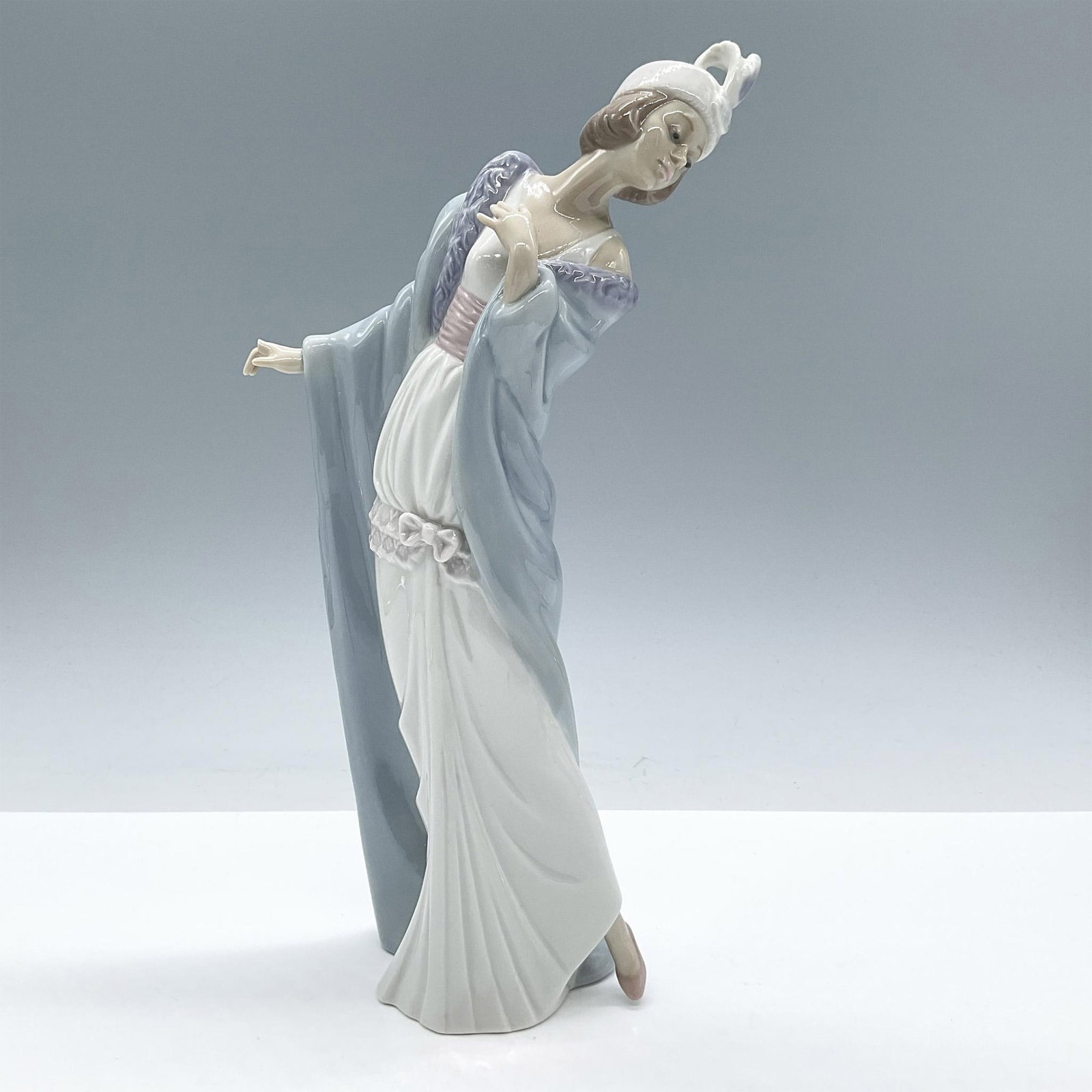 The Flirt 1005789 - Lladro Porcelain Figurine: Glossy porcelain. Features an elegant lady wearing a white gown and blue coat. Lladro backstamp. Includes the original box, dimensions: 8''L x 12.5''W x 6''H. Artist: Joan Coderch Issued: 1991 - 1998 
