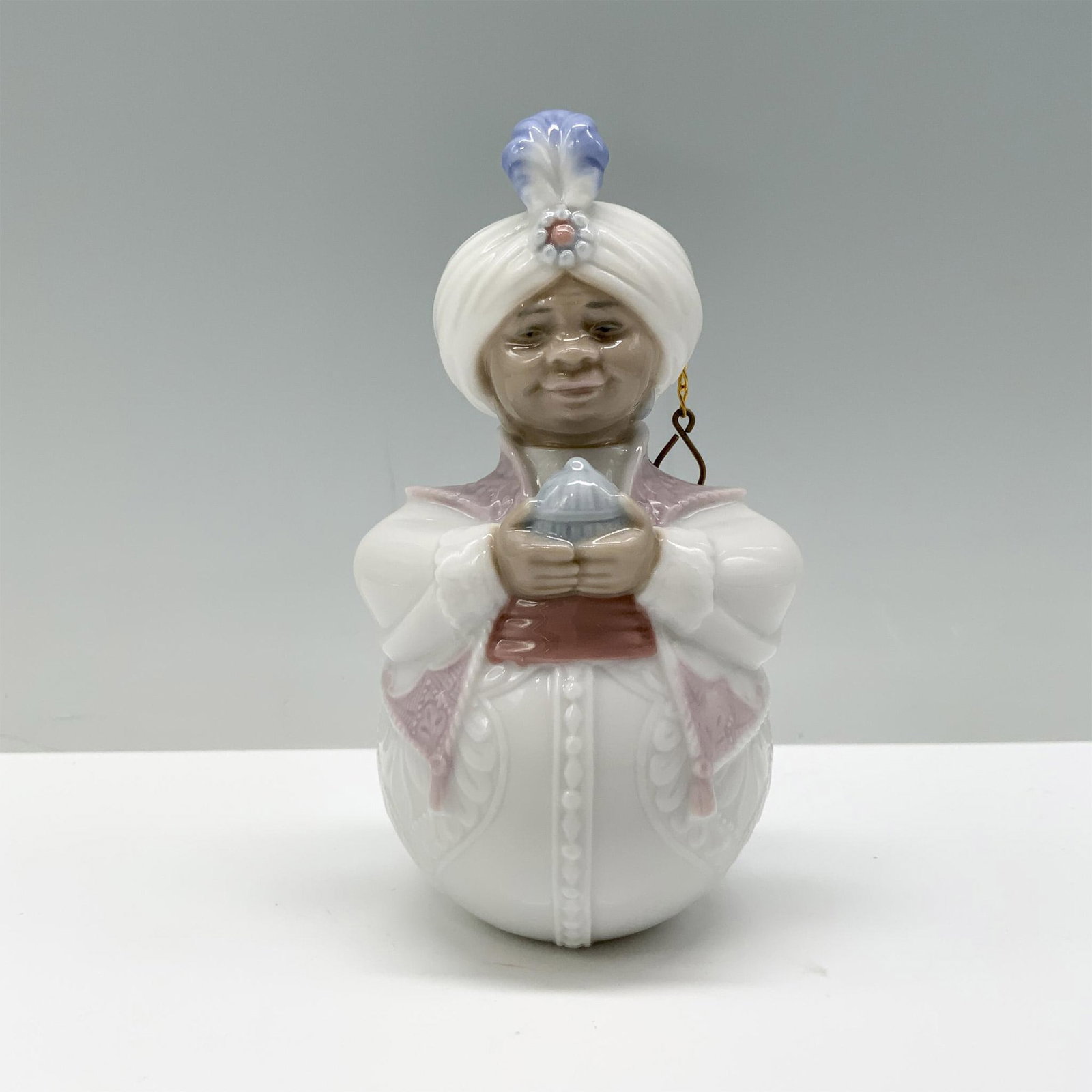 King Balthasar 01006509 - Lladro Porcelain Tree Ornament: King Balthasar, adorned in white and pink attire, holds a gift in his hands with regal grace. Lladro backstamp. This item has its original box which measures: 4.5"L x 4.25"W x 3.75"H. Artist: Jose Puc