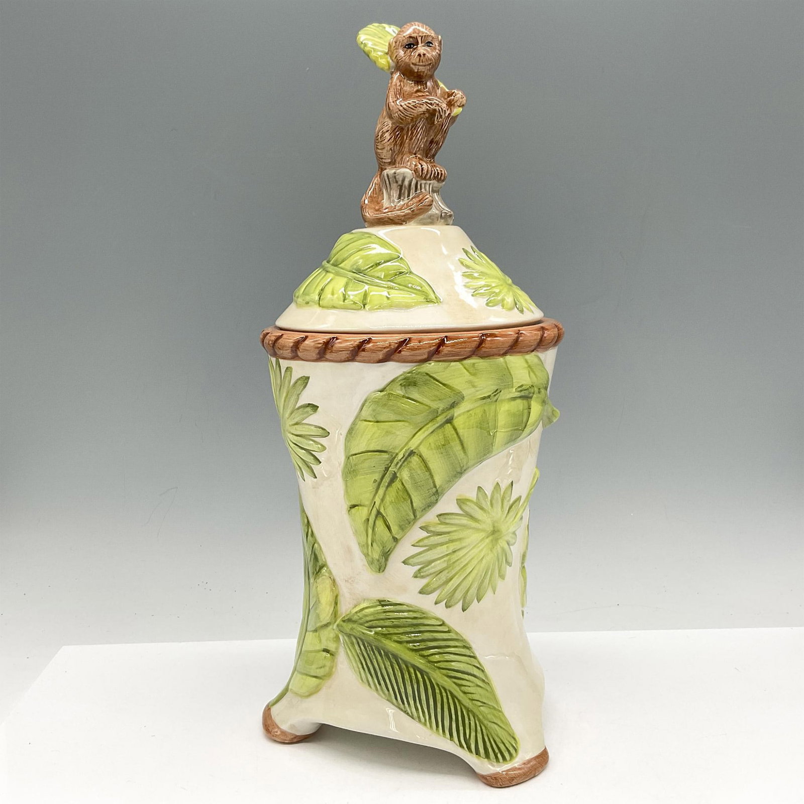American Atelier Porcelain Monkey Canister: Whimsical design of monkey seated as handle with leaf fronds around him and on base. Colored in natural brown and green. American Atelier impressed mark. Issued: 21st century Dimensions: 5.5"L x 5"W x