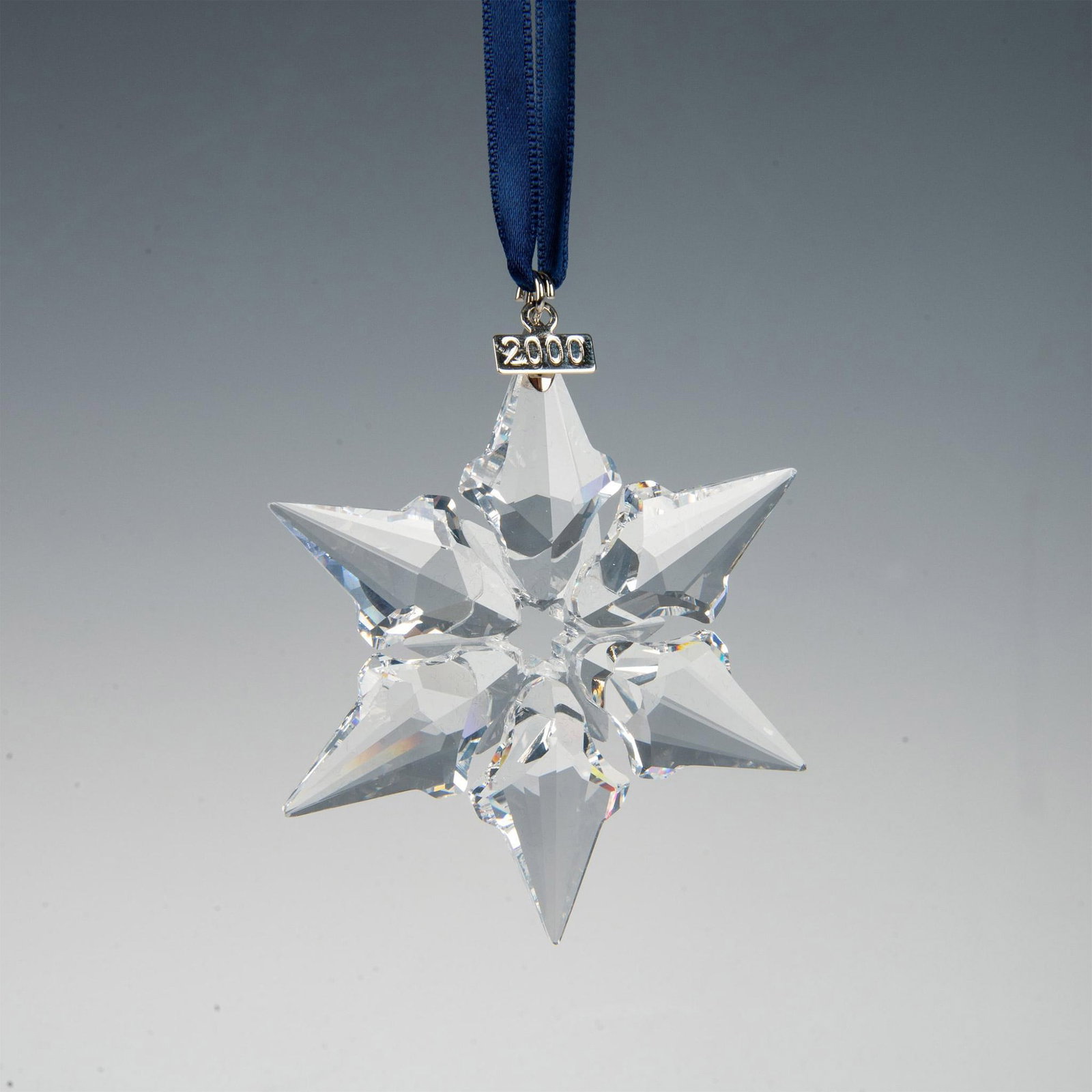 Swarovski Crystal 2000 Christmas Ornament: A clear, faceted six point snowflake adornment featuring a metal tag with 2000 impressed on it, hanging from a navy blue ribbon. Swarovski marking. This item has its original box: 6.5"L x 5.5"W x 1.25