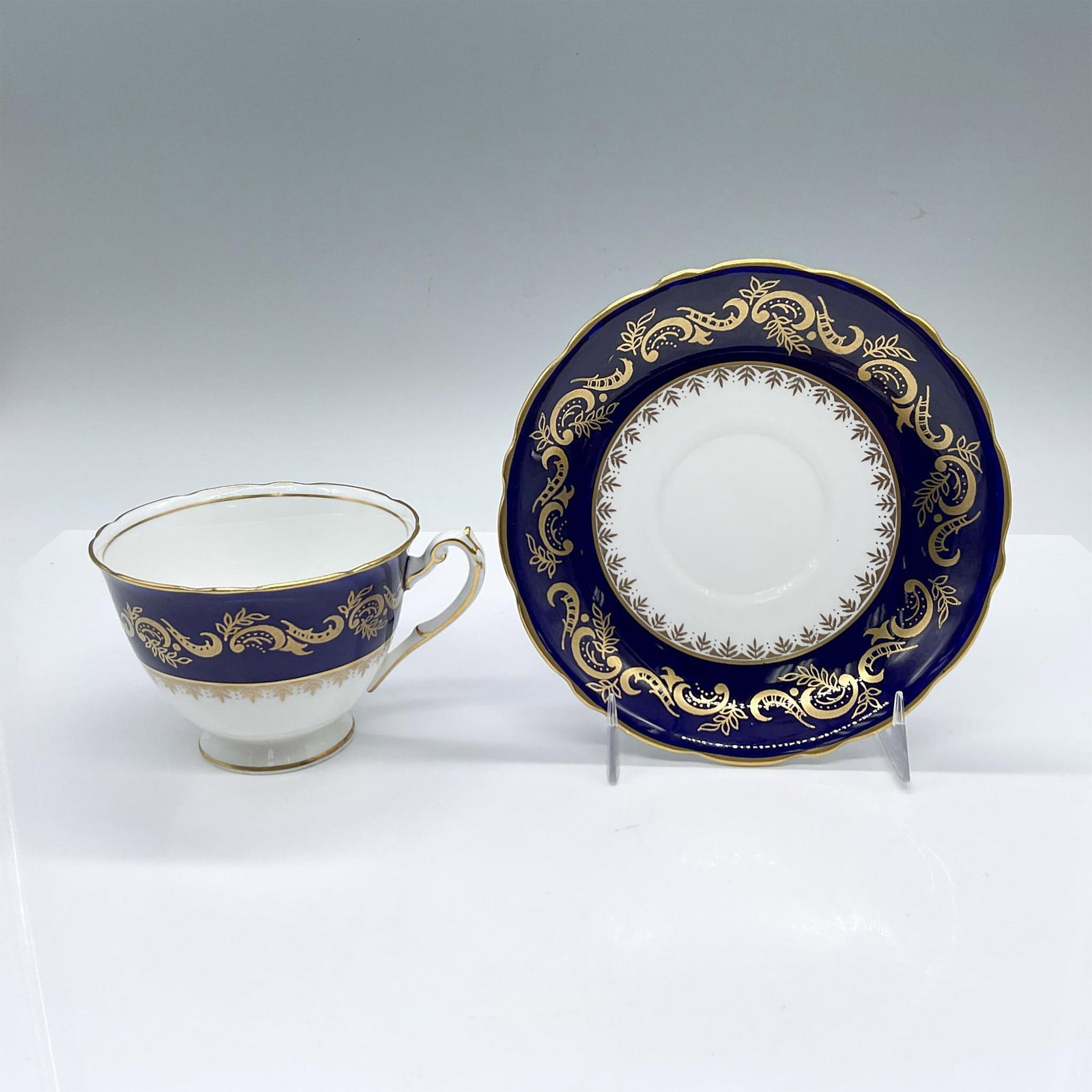 2pc Royal Cauldon Bone China Gilded Blue Tea Cup and Saucer: English white cup and saucer set, decorated in a cobalt-blue band with a gilded acanthus scroll design. Royal Cauldon backstamps. Cup: 4.5"L x 3.75"W x 2.75"H. Saucer: 1"H x 5.75" dia. Issued: 20th ce