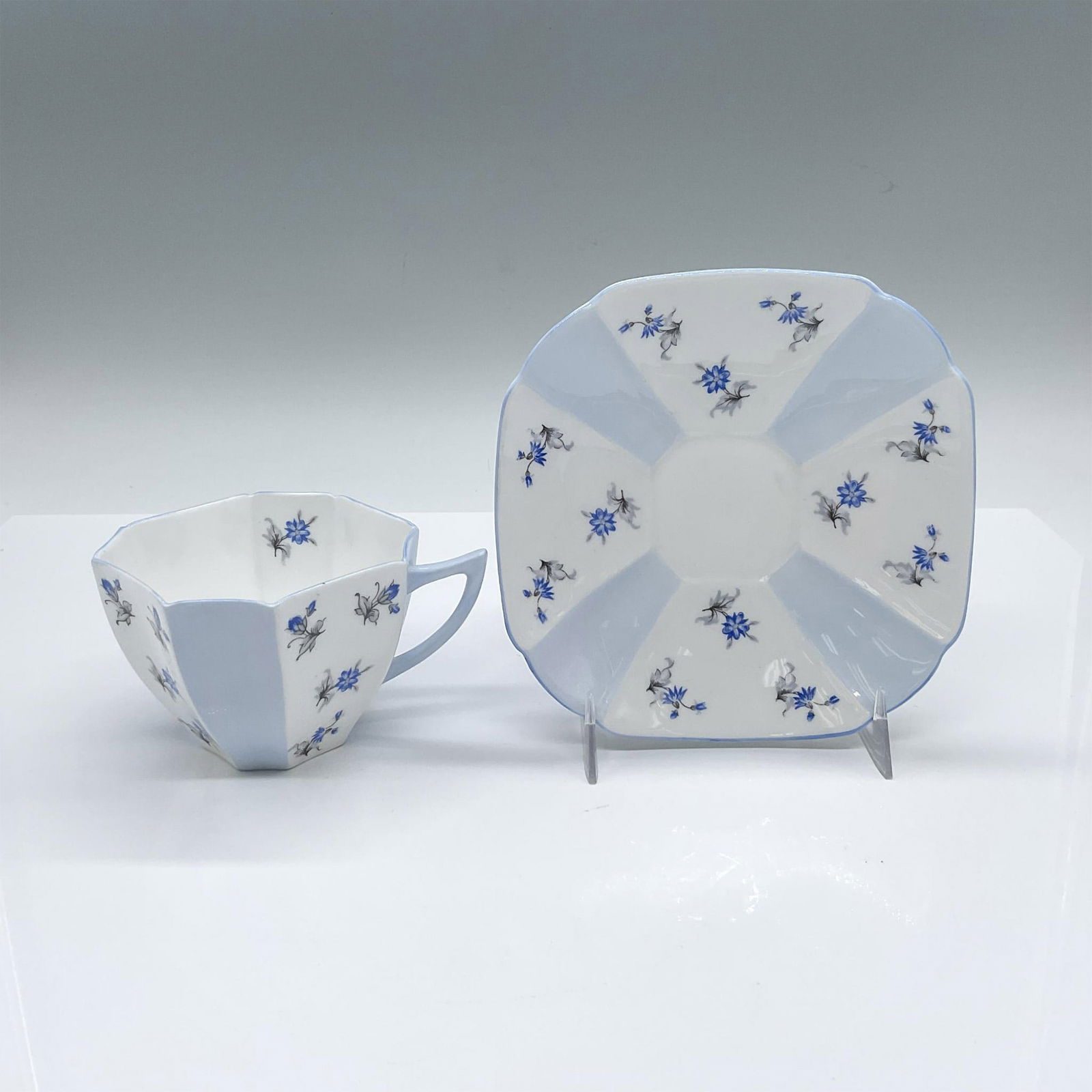2pc Shelley Bone China Queen Anne Cup and Saucer, Blue Charm: English white and blue tea cup and saucer set adorned with blue petal flowers. Blue trim accents. Shelley backstamps. Cup: 4.25"L x 3.5"W x 2.5"H. Saucer: 5.25"L x 5.25"W x 0.75"H. Issued: 20th centur