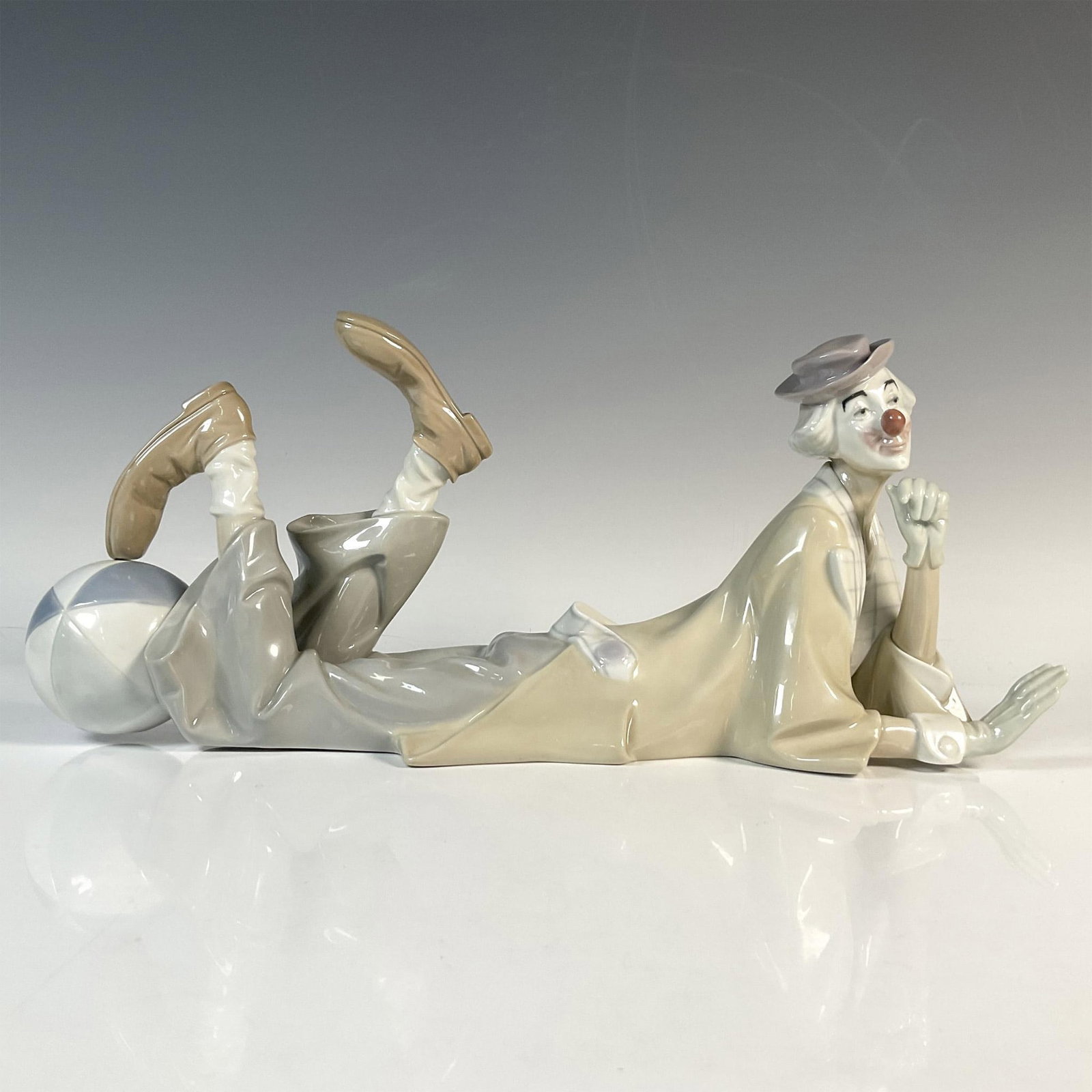 Clown 1004618 - Lladro Porcelain Figurine: A glossy figure of a comedian leisurely laying down with a beach ball by his feet. Lladro backstamp. Artist: Salvador Furio Issued: 1970-2019 Dimensions: 15"L x 5"W x 6.5"H Manufacturer: Lladro Count