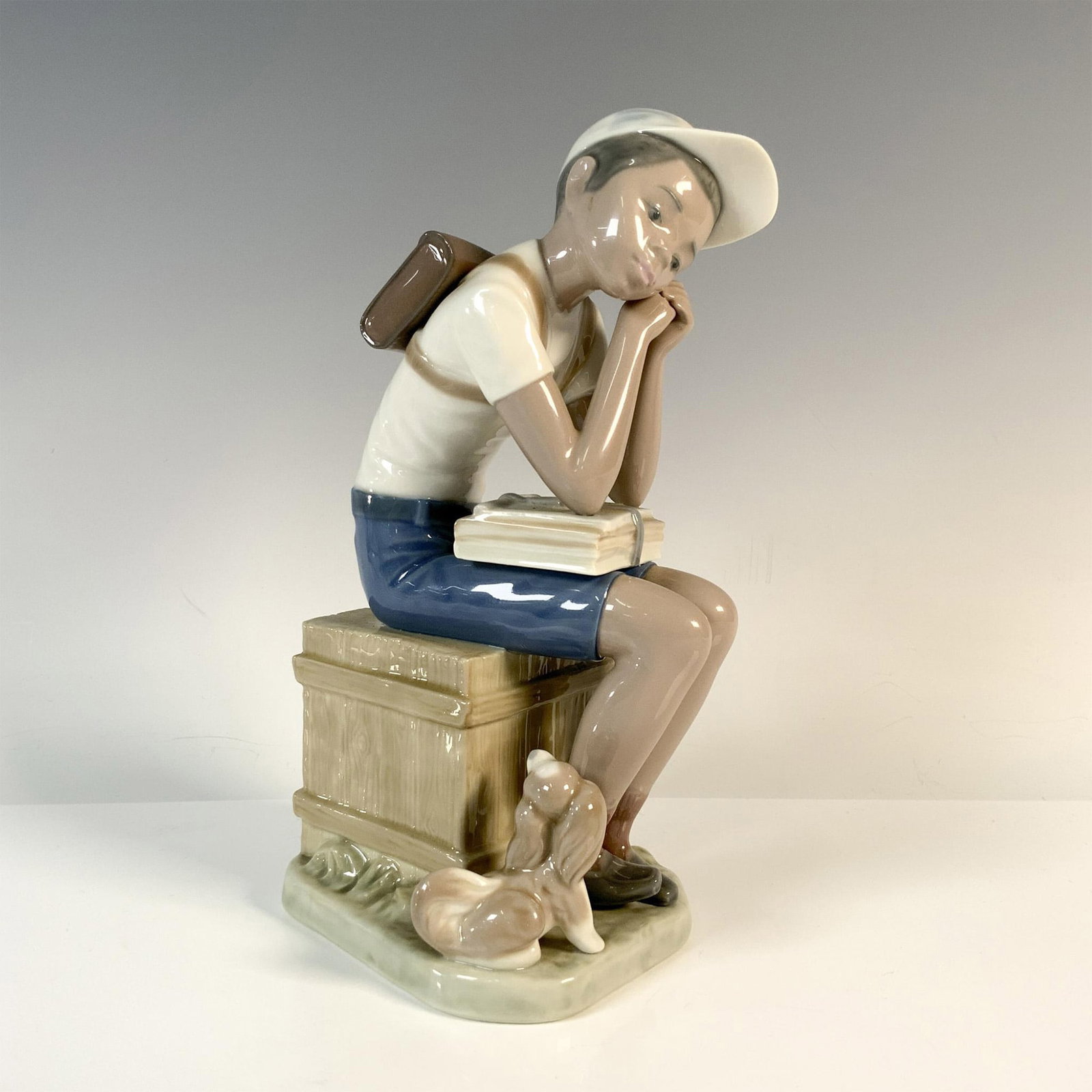 Exam Day 1005250 - Lladro Porcelain Figurine: Depicted wearing white and blue clothes, sitting with his backpack, waiting for his test, accompanied by his little dog. Lladro backstamp. Artist: Francisco Catala Issued: 1984 - 1994 Dimensions: 4''L