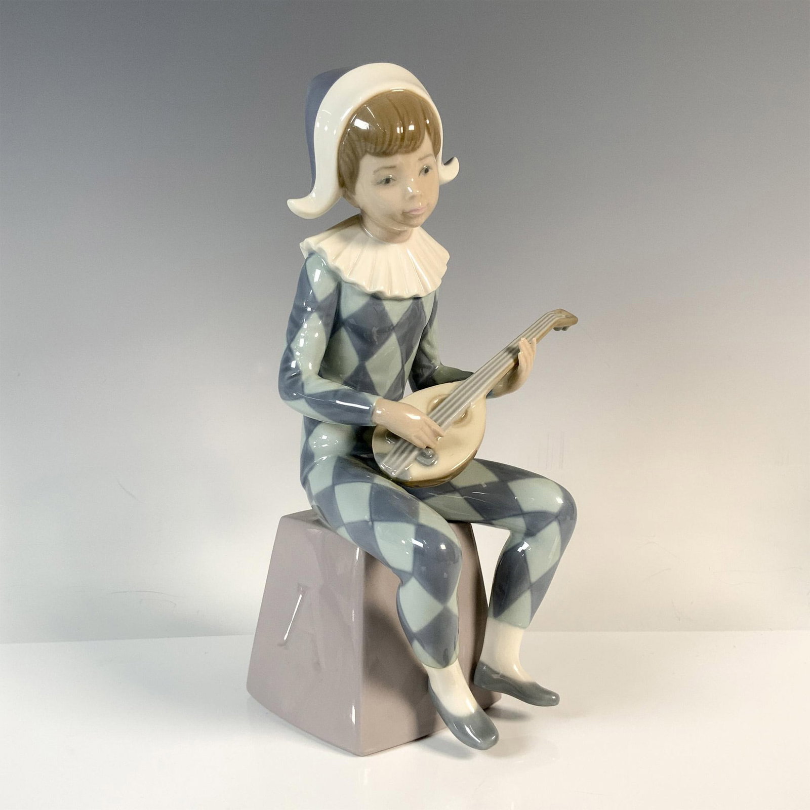 Harlequin A 1005075 - Lladro Porcelain Figurine: Glazed finish. Features a boy sitting wearing blue and green harlequin clothes. Lladro backstamp. Artist: Francisco Catala Issued: 1980 - 1985 Dimensions: 3.75''L x 4''W x 8.75''H Manufacturer: Swarov