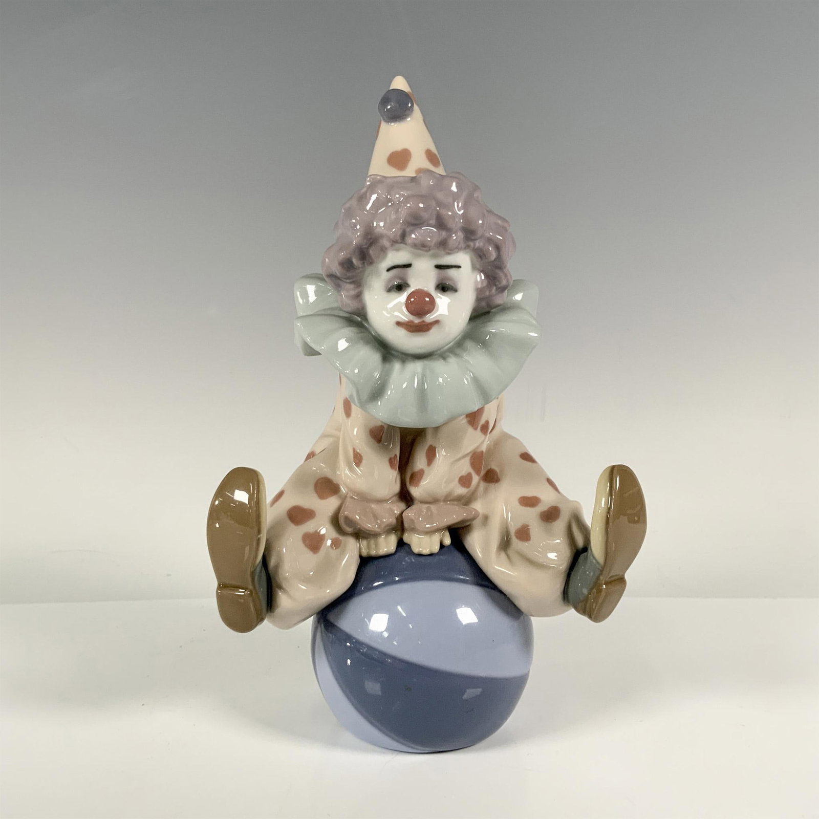 Having A Ball 1005813 - Lladro Porcelain Figurine: Features a playful clown playing on a blue ball, dressed on a red suit with hearts. Lladro backstamp. Artist: Antonio Ramos Issued: 1991 - 2018 Dimensions: 3''L x 4.25''W x 7''H Manufacturer: Lladro