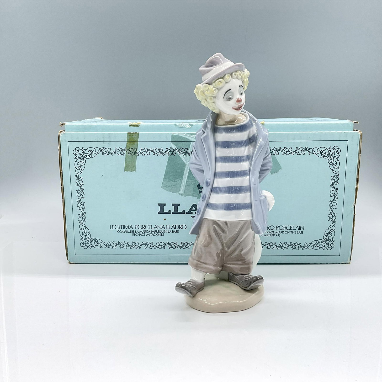 Little Traveler 1007602 - Lladro Porcelain Figurine: Glossy, hand made porcelain, crafted in pale pastel hues of blue and brown. Depicts a clown in a contemplative pose, carrying a bag. Includes the original box, dimensions: 12.5''L x 7.5''W x 6''H. Lla