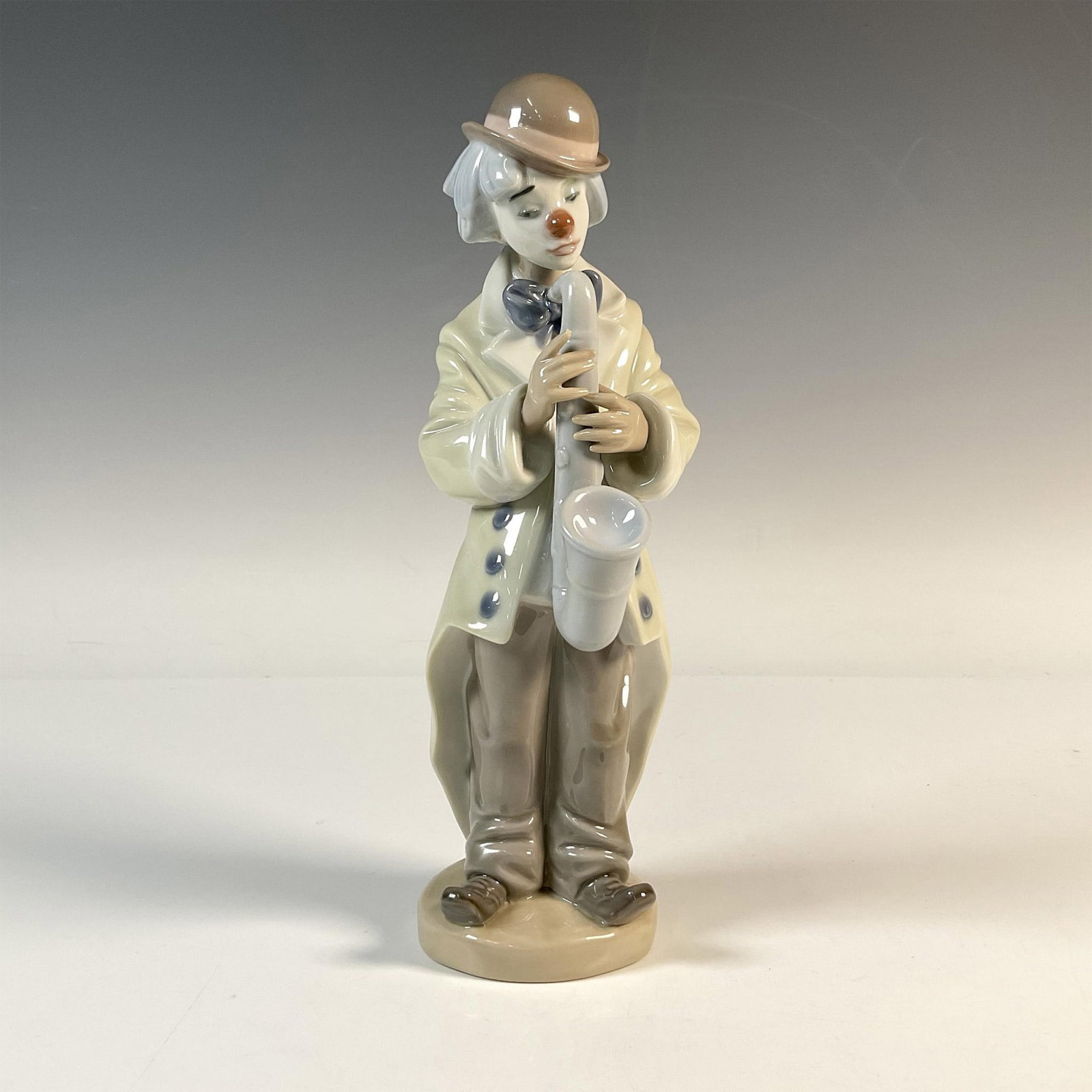 Sad Sax 1005471 - Lladro Porcelain Figurine: A glossy figure depicting a clown in an oversized jacket with a gloomy expression, holding a saxophone. Lladro backstamp. Artist: Francisco Catala Issued: 1988-2018 Dimensions: 3"L x 4"W x 9"H Manufac