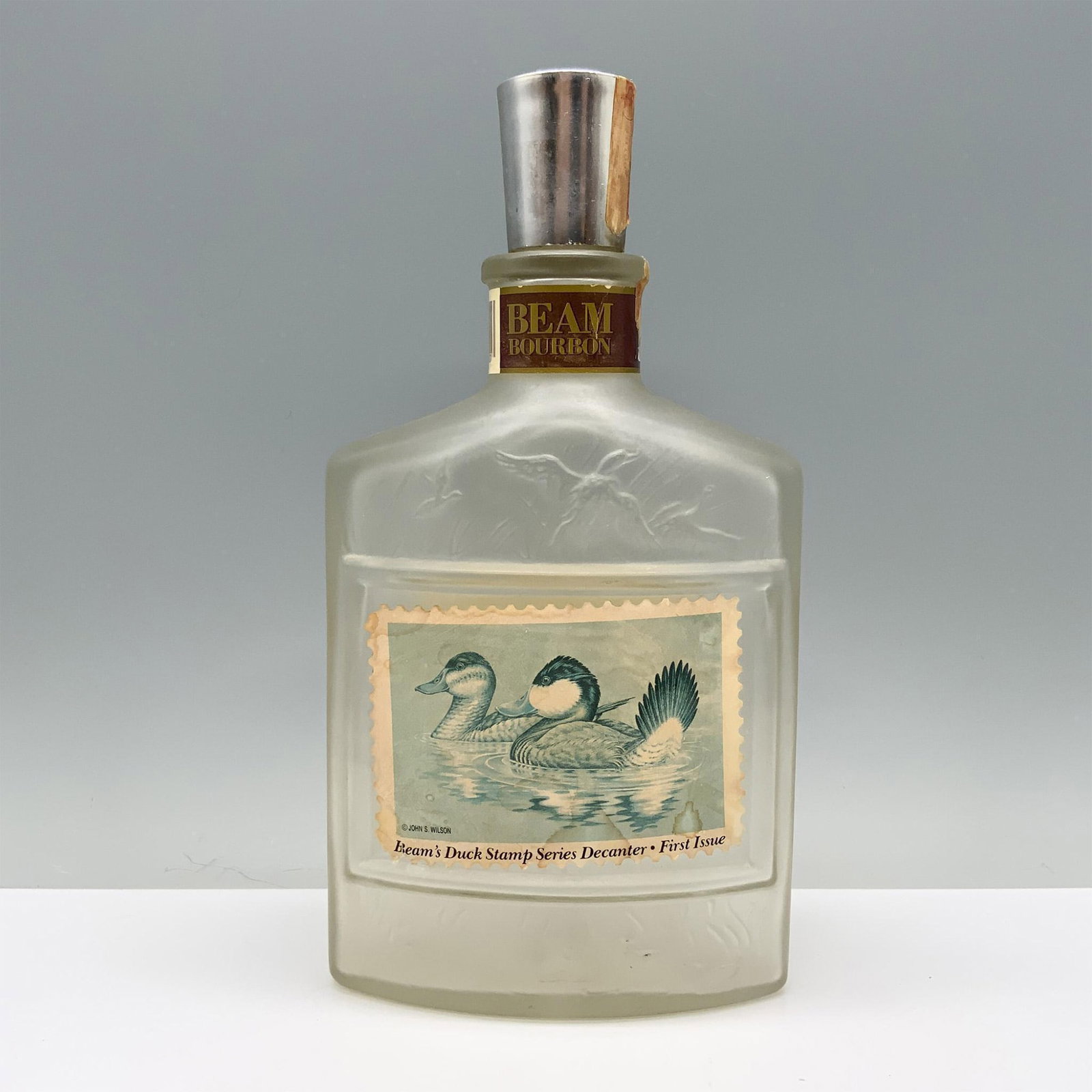 Beams Duck Stamp Series Decanter, Ruddy Ducks: A clear decanter intricately decorated with raised images of ducks in flight from tall grass, centered around a prominent stamp of a duck couple swimming, with a list of other editions on the back. Be