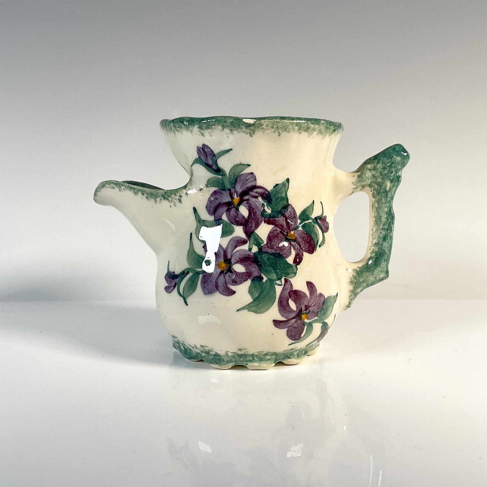 Cash Family Art Pottery Creamer, Violets: A cream-colored creamer featuring green accent borders and delicate artwork showcasing violets, complete with three convenient pour spots at its top. Marked Cash Family. Dimensions: 5"L x 2.75"W x 3.2