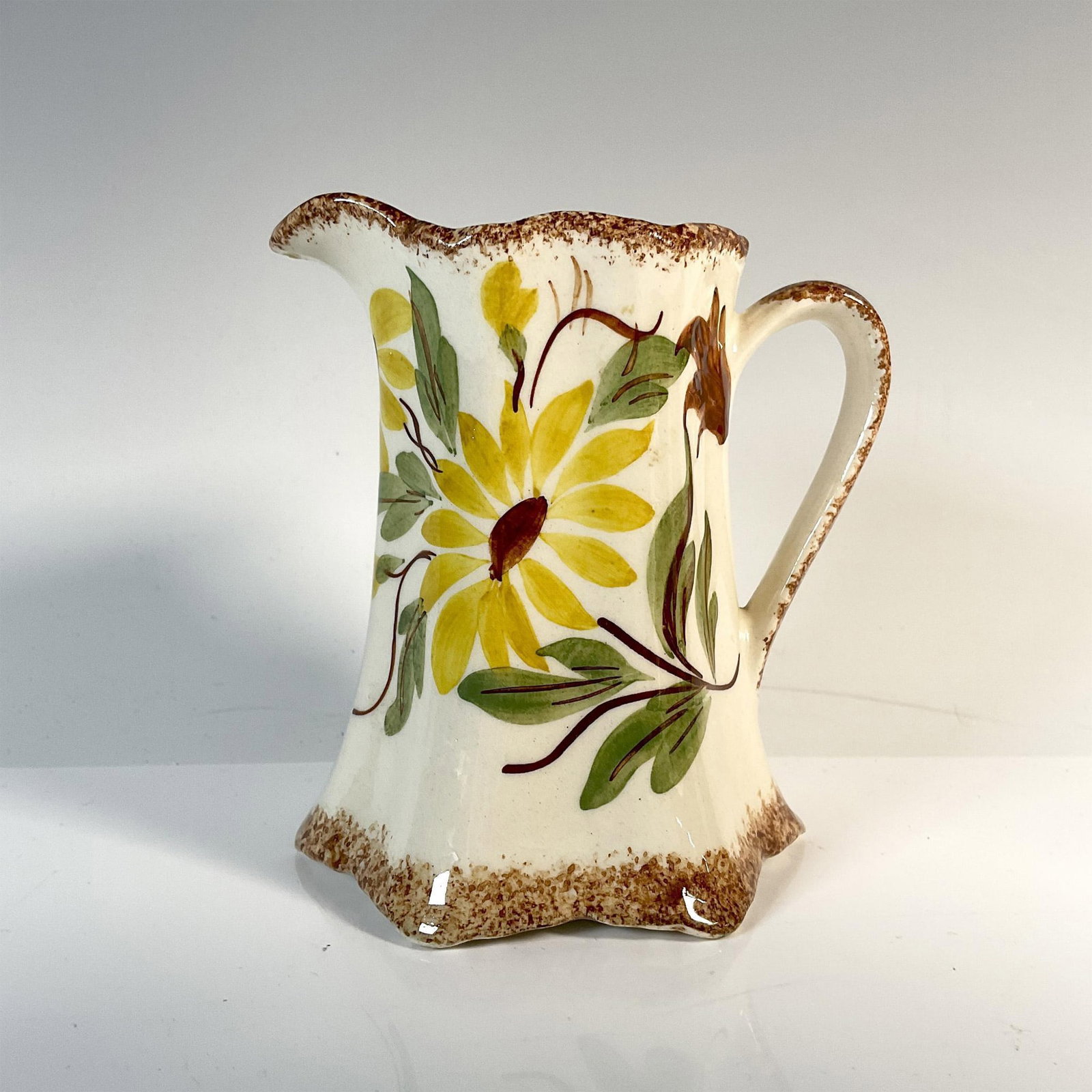 Cash Family Art Pottery Creamer, Sunflowers: A cream-colored creamer with brown accent borders, adorned with charming artwork depicting a sunflower and its foliage. Marked Cash Family. Dimensions: 3.75"L x 3"W x 4"H Manufacturer: Cash Family Cou