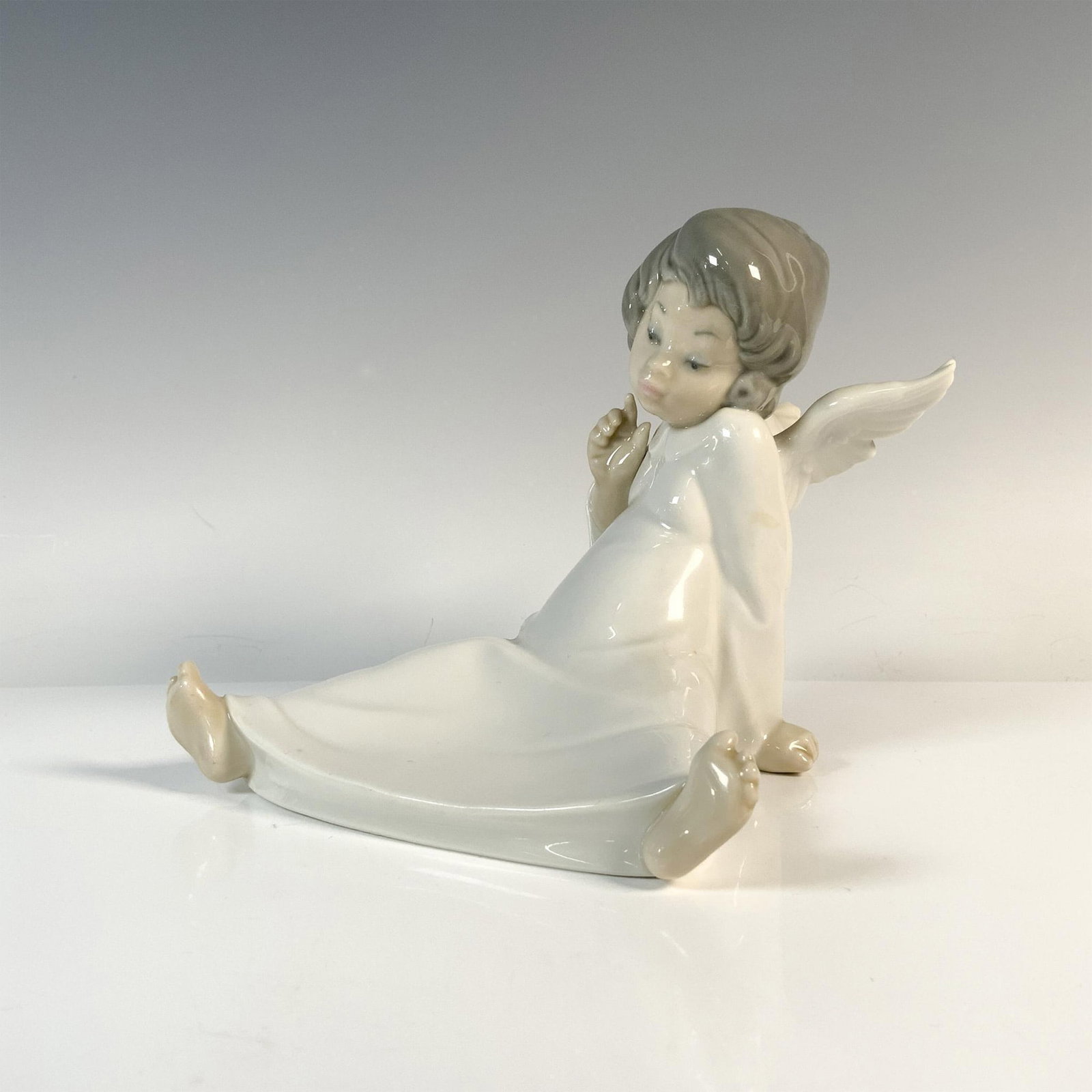 Cherub Wondering 1004962 - Lladro Porcelain Figurine: Glazed figurine of a seated angel. Lladro backstamp. Artist: Salvador Debon Issued: 1977-2017 Dimensions: 6.5"L x 6"W x 5.5"H Manufacturer: Lladro Country of Origin: Spain