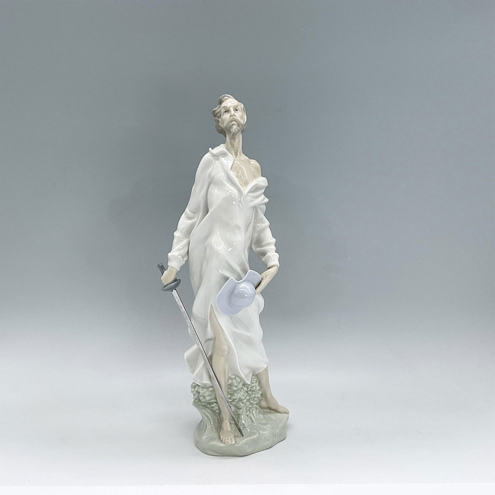 Don Quixote - Nao by Lladro Porcelain Figurine: Glazed porcelain figure of Don Quixote standing in a white nightshirt holding a sword in one hand and his blue hat in the other. Nao backstamp. Dimensions: 4.75"L x 3.25"W x 1.5"H Manufacturer: Nao by