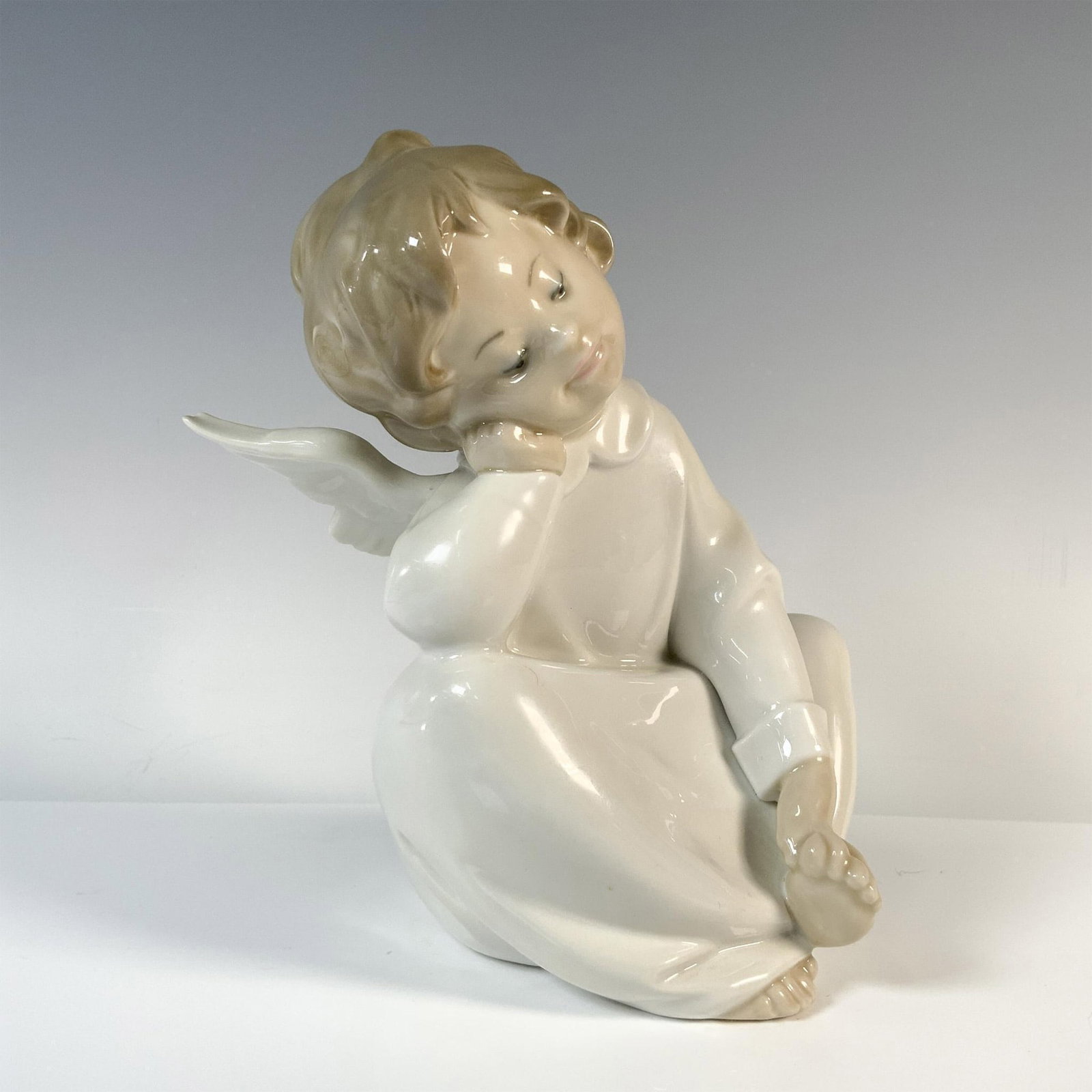 Dreaming Cherub 1004961 - Lladro Porcelain Figurine: A figurine of a young angel child, seated in peaceful contemplation with his head resting gently on hand, adorned in a flowing white robe, and majestic open wings. Lladro backstamp. Artist: Salvador D