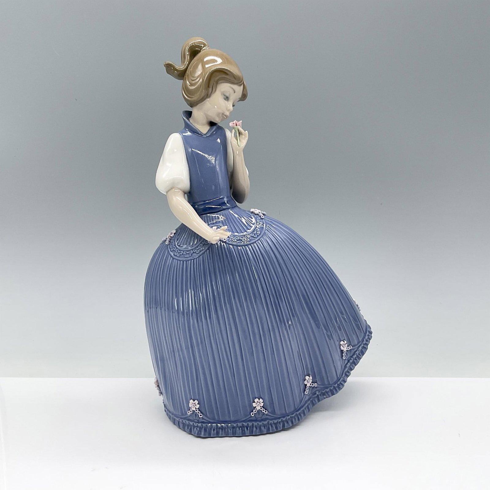 Girl Bluish Dress Flower 1005119 - Lladro Porcelain Figurine: Beautiful glossy figurine of young girl in a blue dress with rippled skirt adorned with tiny delicate flowers. Lladro backstamp. Artist: Jose Puche Issued: 1982 Dimensions: 7.25"L x 6.5"W x 11"H Manuf