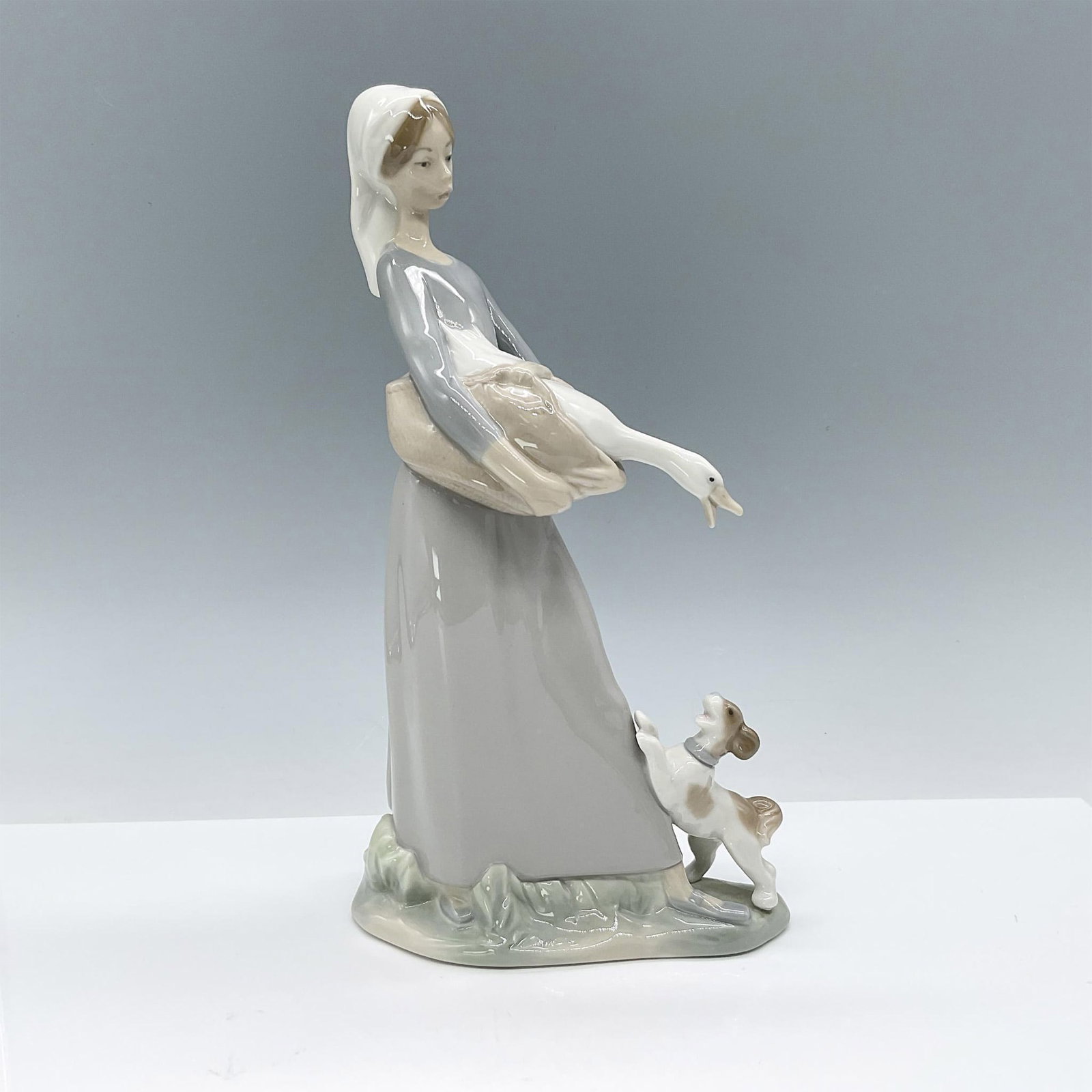 Girl with Goose and Dog 1004866 - Lladro Porcelain Figurine: Glossy finished figural girl carrying basket with goose, dog barking at her feet. Colored in tan, grey and white. Lladro backstamp. Artist: Fulgencio Garcia Issued: 1970 - 1992 Dimensions: 6"L x 3.5"W