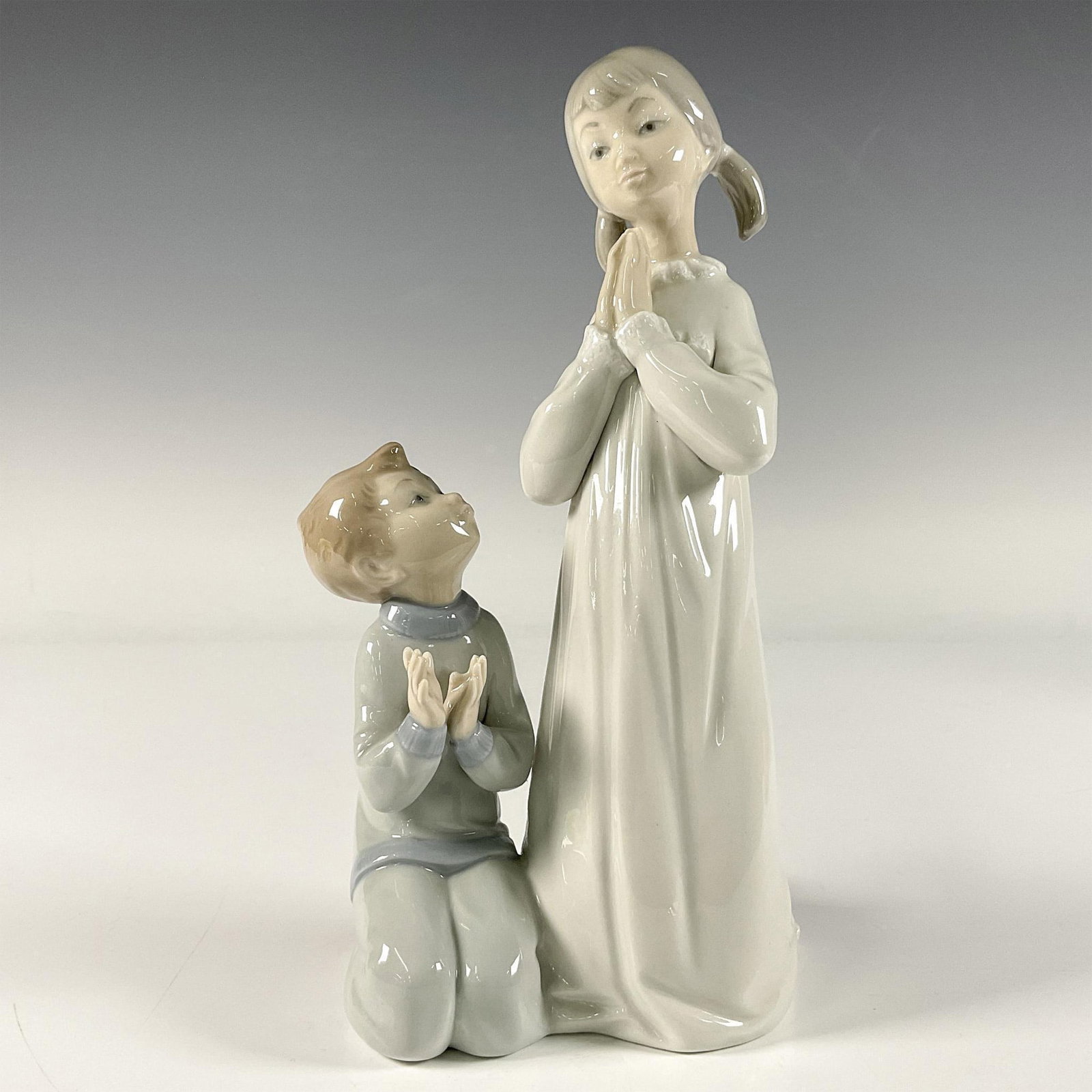 Teaching to Pray 1004779 - Lladro Porcelain Figurine: A glossy figure depicting an older sister instructing her younger brother on how to pray. Lladro backstamp. Artist: Vincente Martinez Issued: 1971-1997 Dimensions: 4.5"L x 4"W x 8.75"H Manufacturer: L