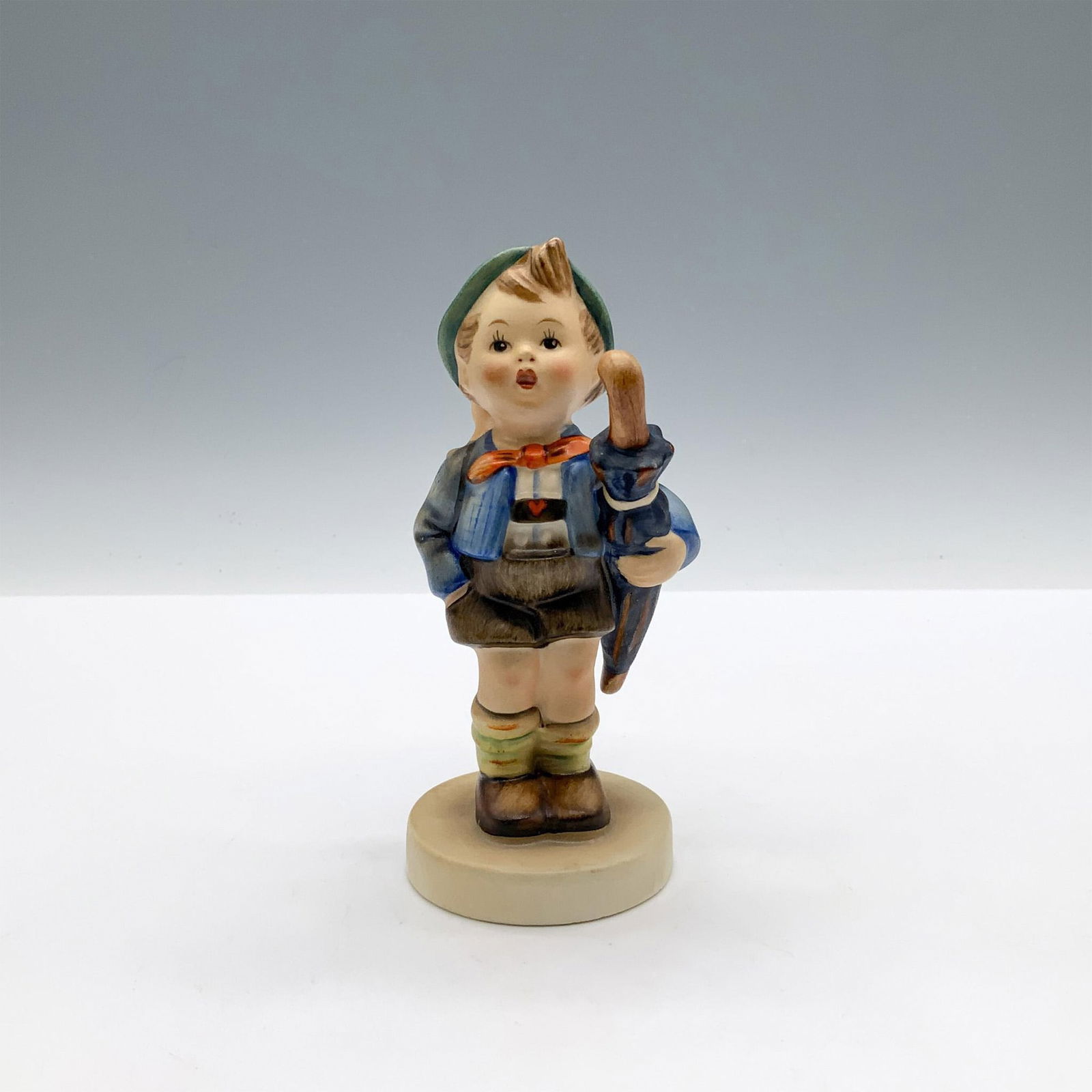 Goebel Hummel Porcelain Figurine, Home From Market: A matte hand painted figure of a boy carrying an umbrella under his arm, and a pig inside a basket on his back. Goebel backstamp. M.I. Hummel impressed. Issued: 1948-2007 Dimensions: 2"L x 2.5"W x 4.7