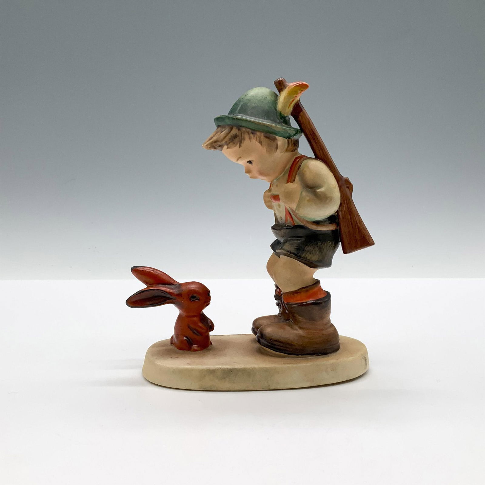 Goebel Hummel Porcelain Figurine, Sensitive Hunter HUM6/0: A matte figure of boy carrying a rifle over his shoulder while he is having a conversation with a small red rabbit. Goebel backstamp. M.I. Hummel impressed. Issued: c. 1935 Dimensions: 4"L x 2"W x 4.7