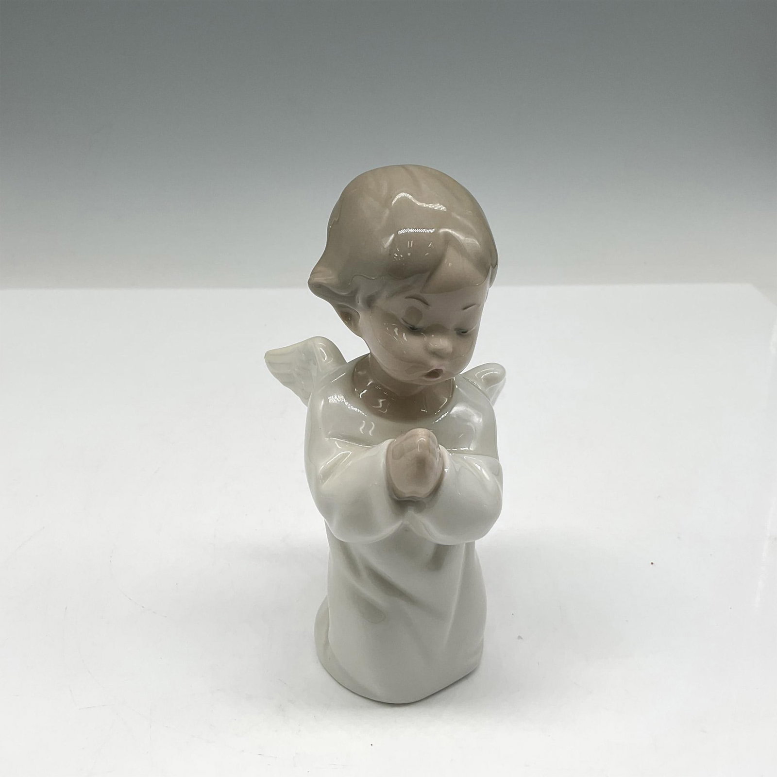 Angel Praying 1004538 - Lladro Porcelain Figurine: A figurine of a young angel child in serene prayer, adorned in a pristine white robe with graceful, open wings. Lladro backstamp. This item has its original box: 5.25"L x 8"W x 5"H. Artist: Fulgencio