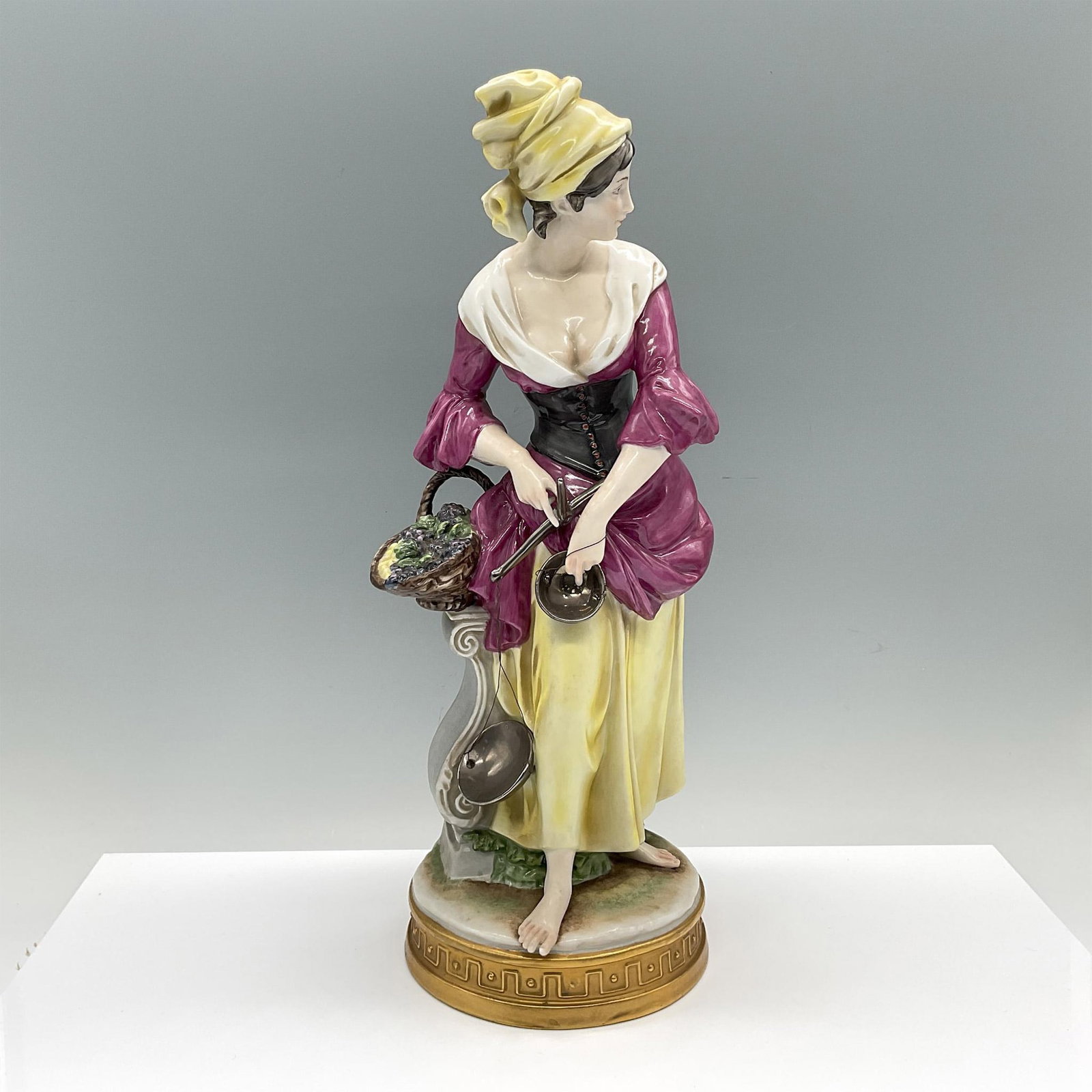 Capodimonte Style Porcelain Peasant Figurine: A painted figure of a maiden in a purple-yellow dress holding a silver-tone scale. She is depicted standing beside a pedestal with grape-filled basket on top. The figure is an allegory of abundance. A
